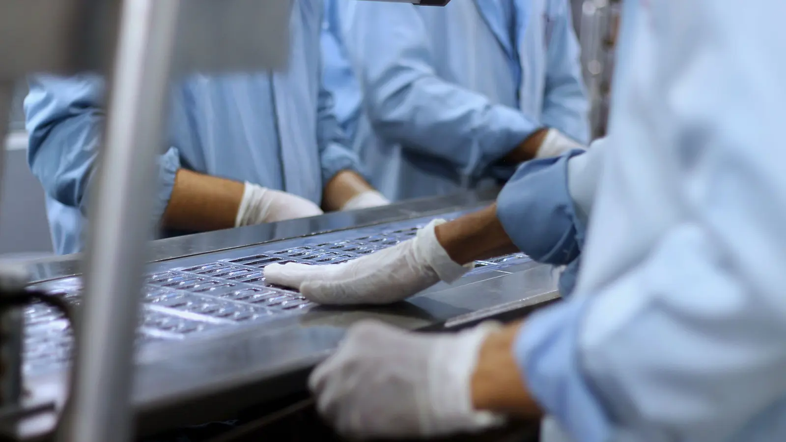 Mani con guanti bianchi e camici azzurri controllano blister su un piano in acciaio