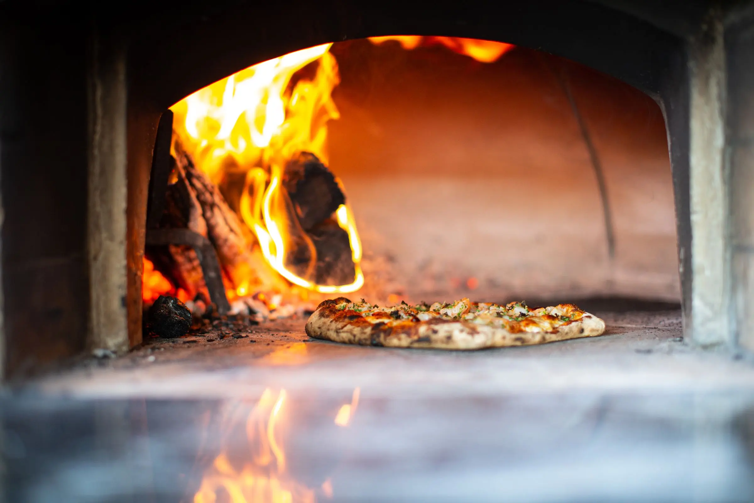 Una pizza nel forno a legna, con la fiamma viva sulla sinistra e il piano di pietra in primo piano