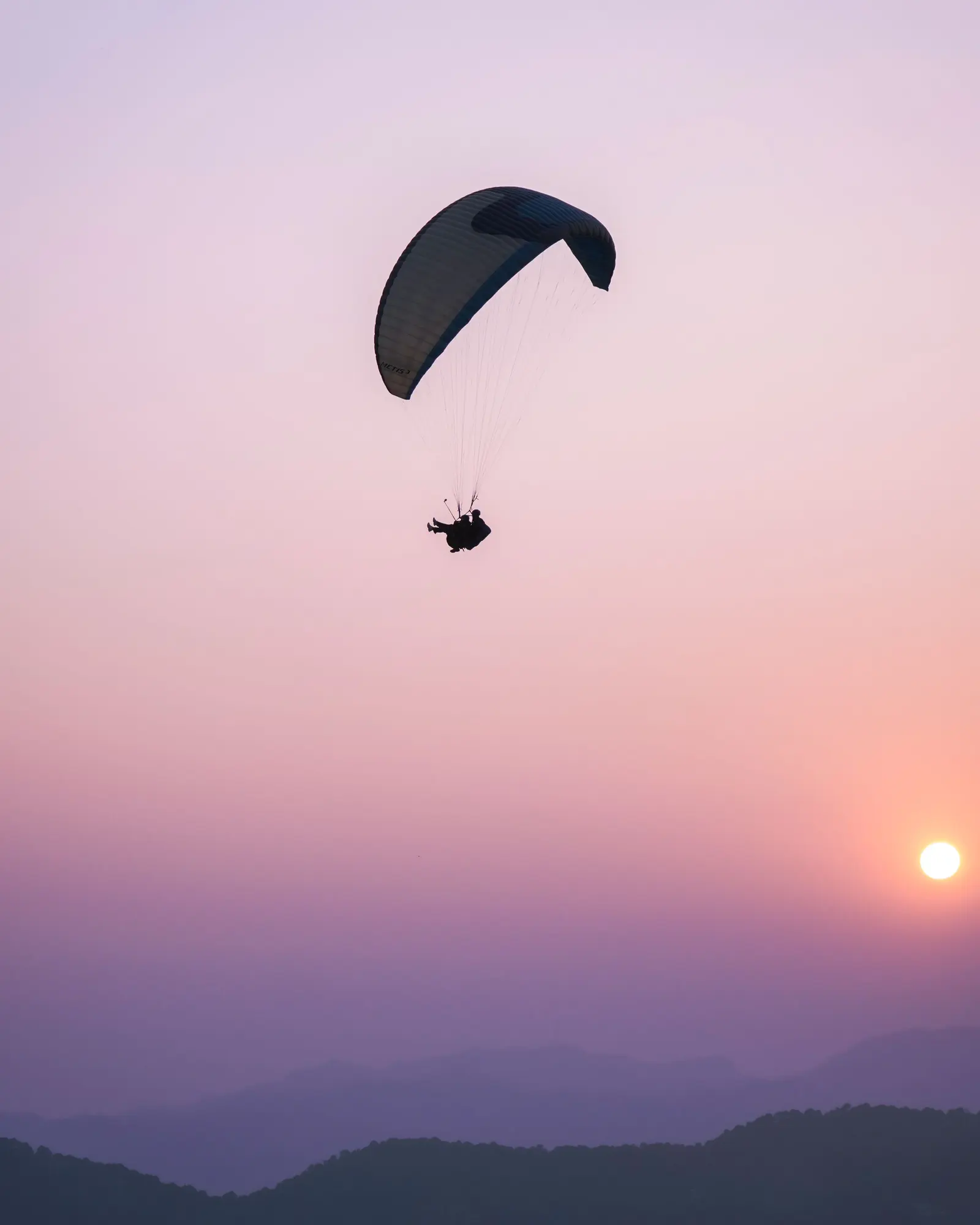 Un parapendio in controluce nel cielo rosa del tramonto, il sole basso sopra le montagne