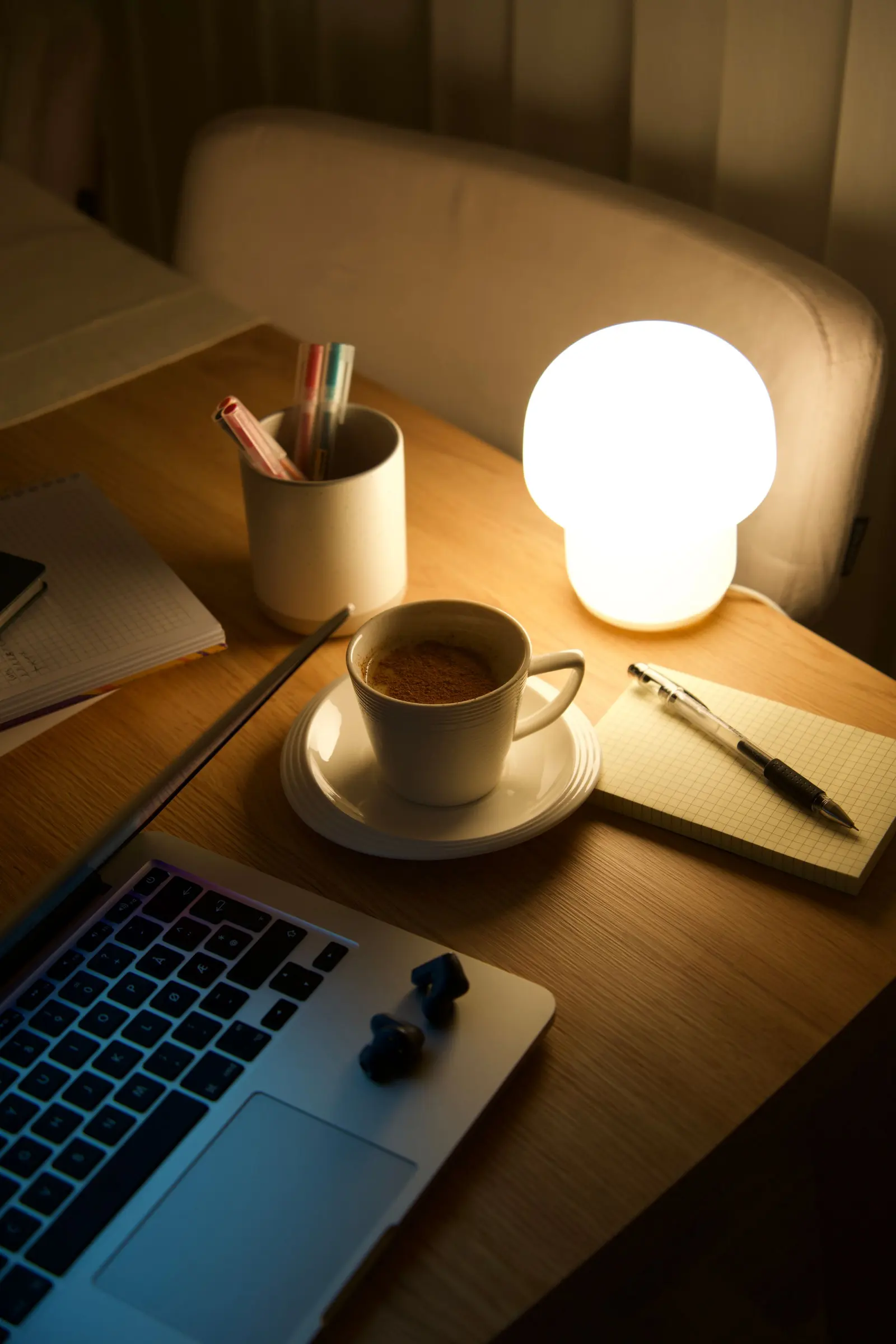 Scrivania di sera con una lampada accesa, il portatile aperto, una tazza di caffè e un taccuino