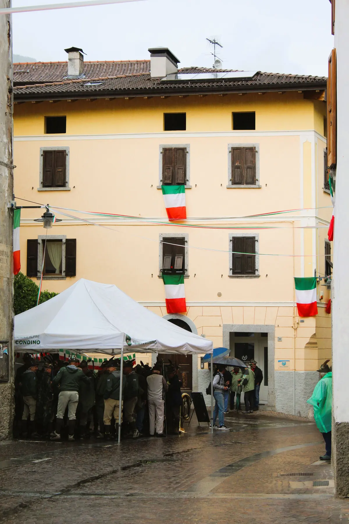 Una festa di paese in una piazzetta, con un gazebo bianco e le bandiere tricolori alle finestre di una casa gialla