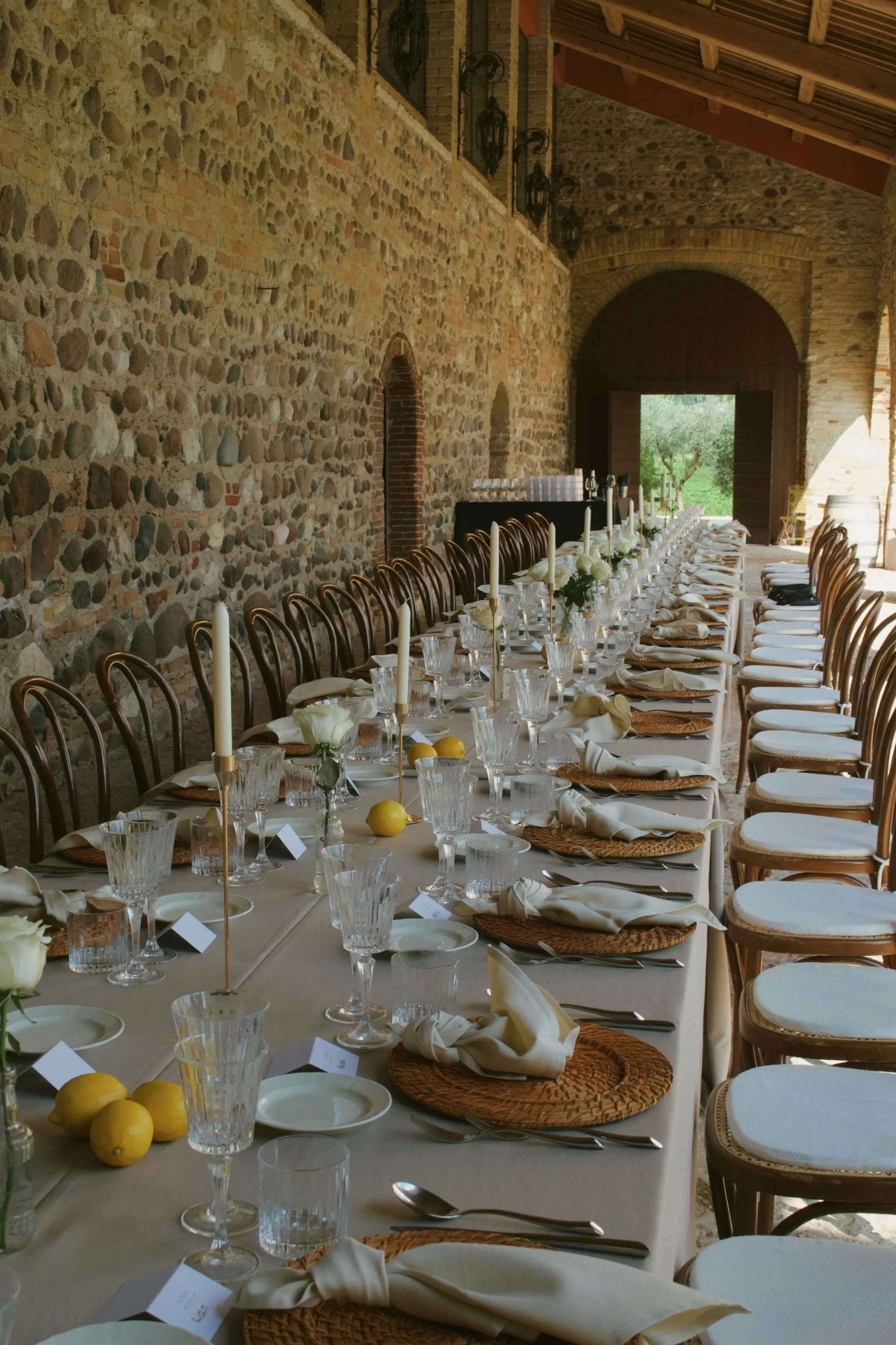 Una lunga tavolata sotto un portico di pietra: tovaglia chiara, sottopiatti di paglia, calici di cristallo, candele e limoni sparsi