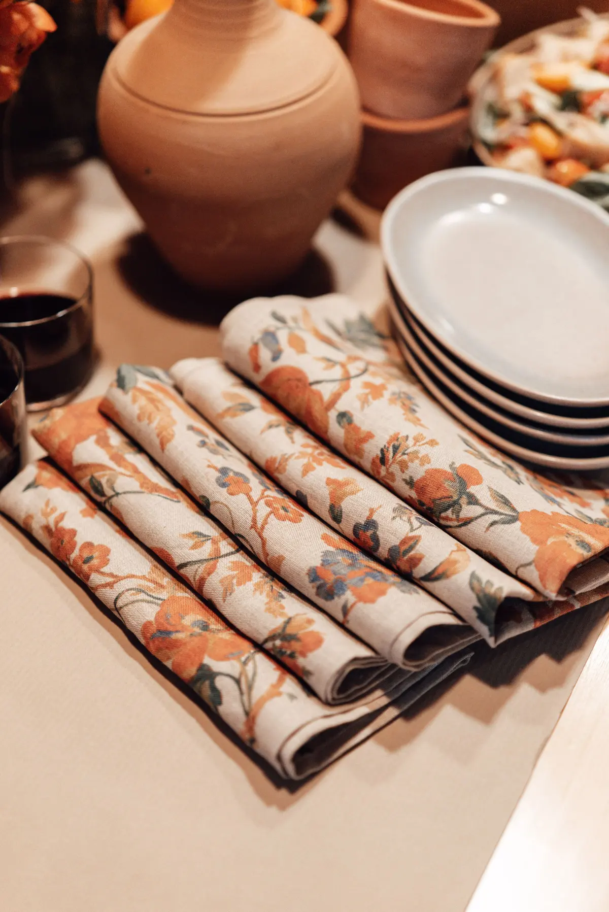 Tovaglioli con una stampa a fiori arancio piegati accanto a piatti chiari e a un vaso di terracotta