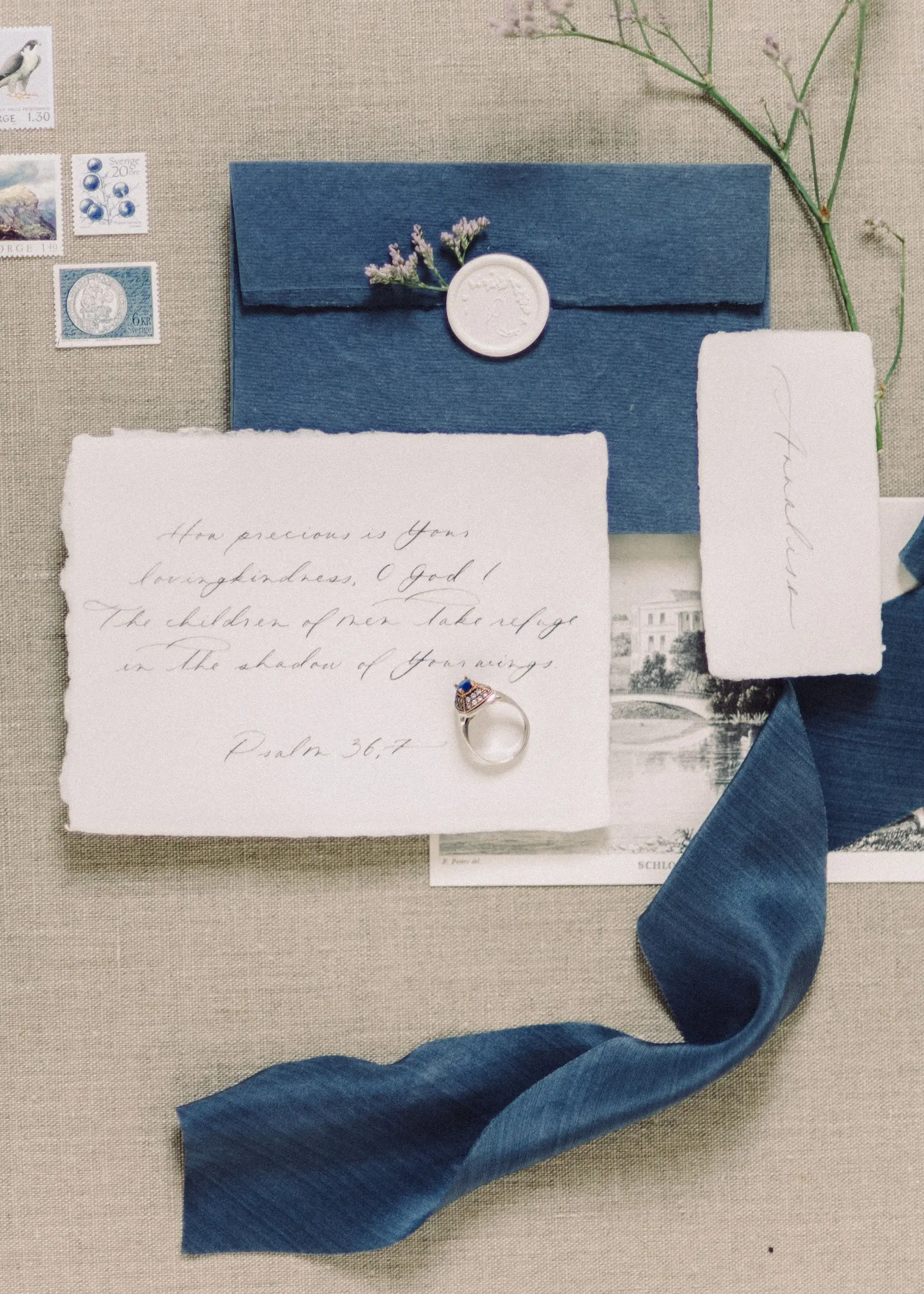 Invito su carta di cotone sfrangiata accanto a una busta blu chiusa da un sigillo bianco, un nastro di seta blu e due rametti su un fondo di lino