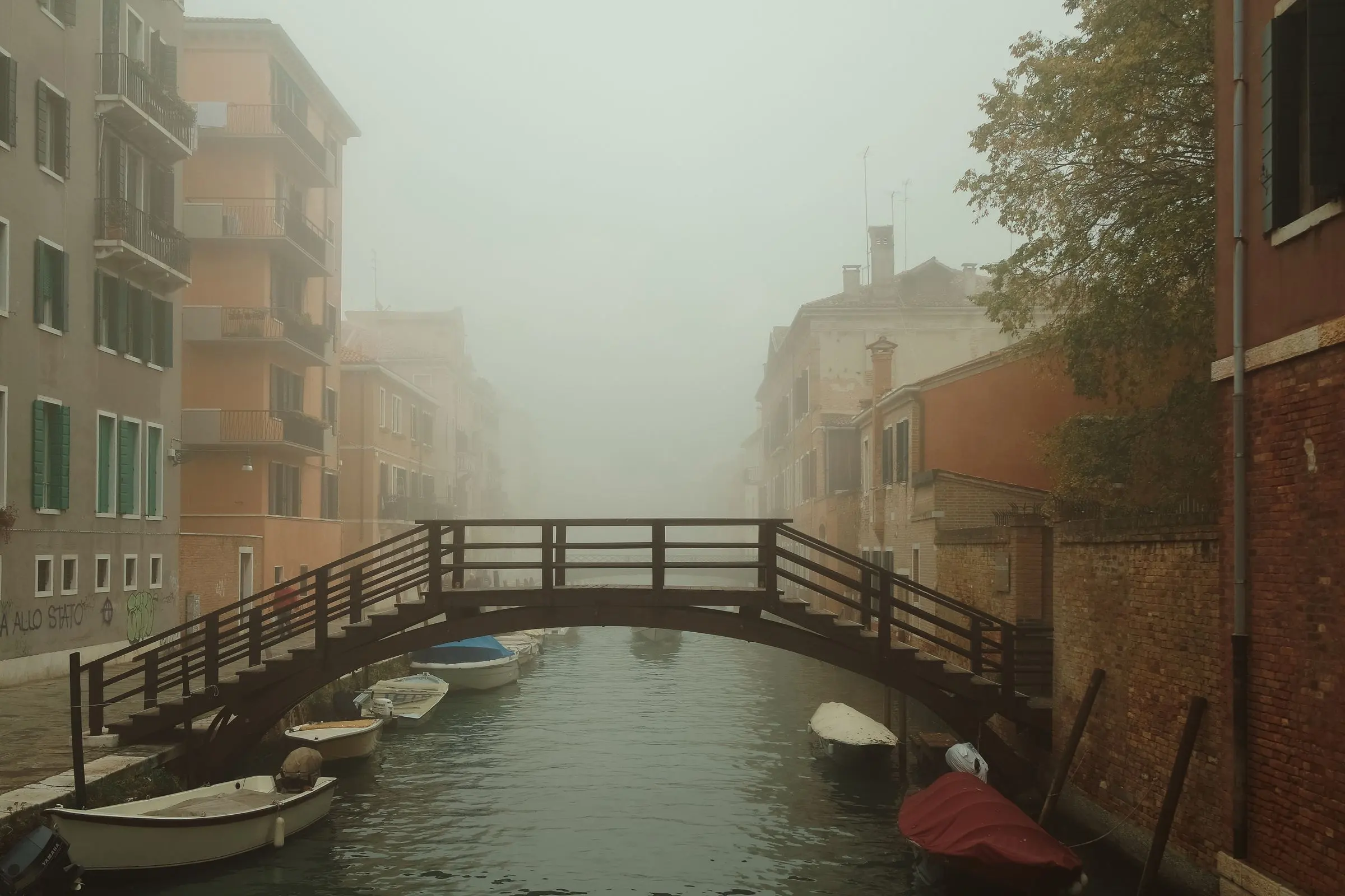Un canale di Venezia nella nebbia del mattino, un ponte di legno e alcune barche ormeggiate tra le case