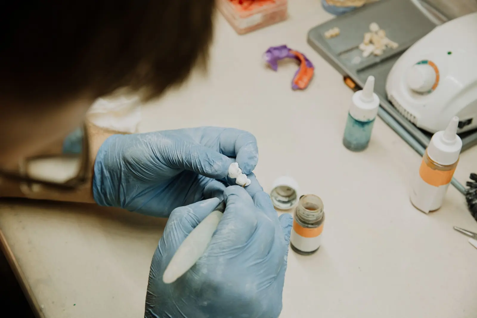 Un odontotecnico rifinisce a mano un piccolo elemento al banco, con i flaconi di colore accanto