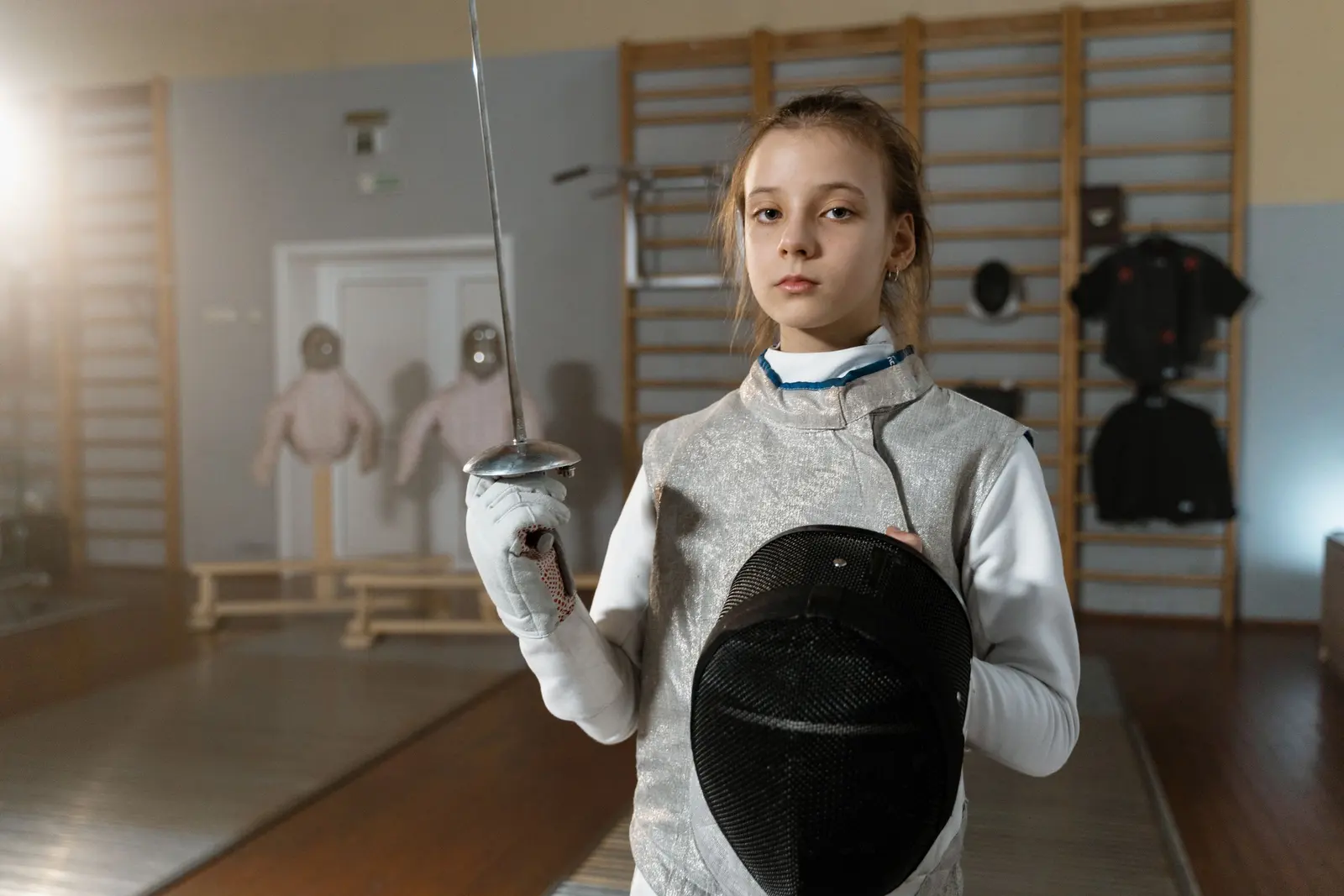 Ragazzina in divisa da scherma con la maschera sotto il braccio e il fioretto in mano