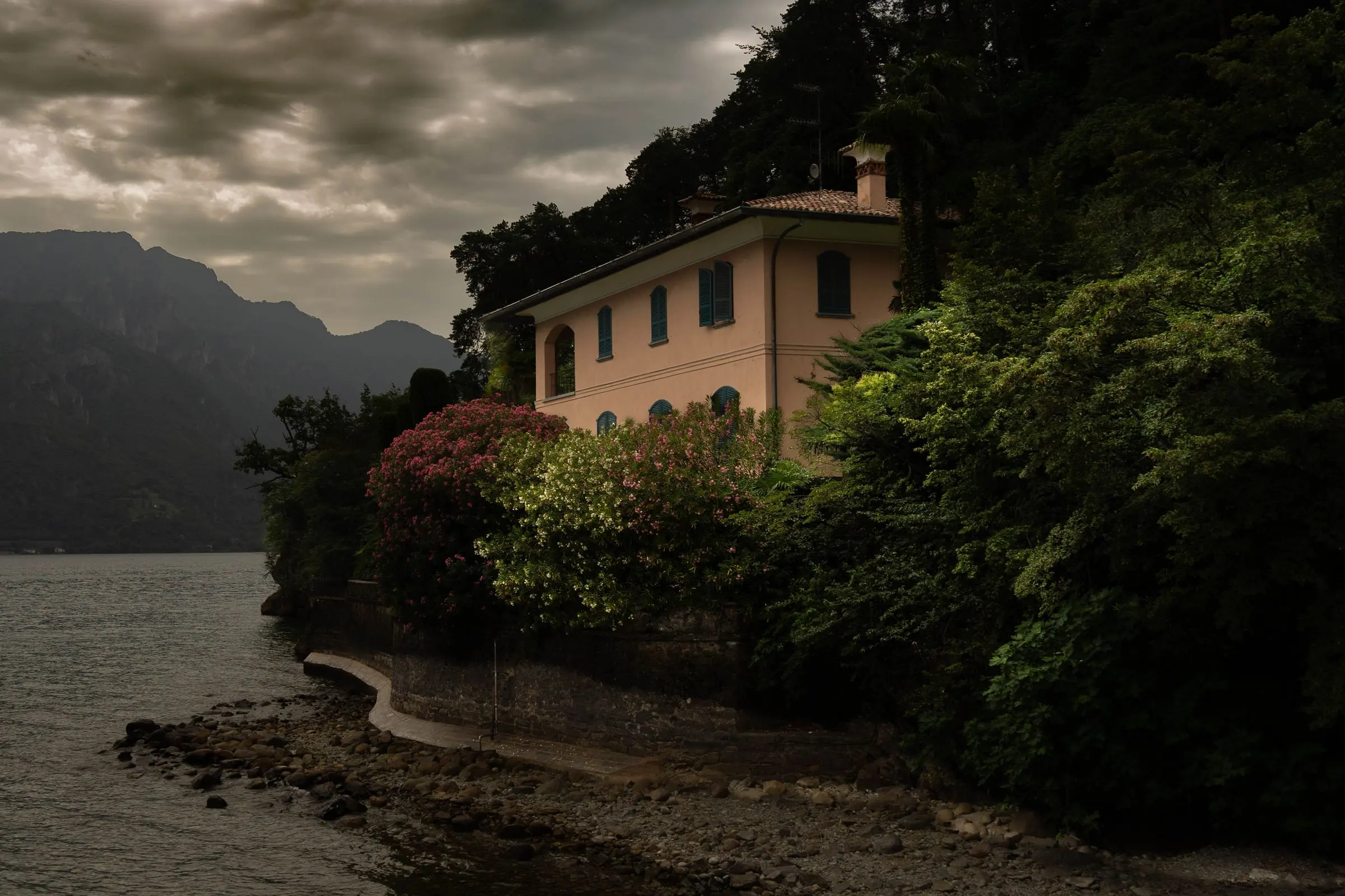 Una villa color ocra tra gli alberi sulla riva del lago, sotto un cielo coperto, con le montagne sullo sfondo