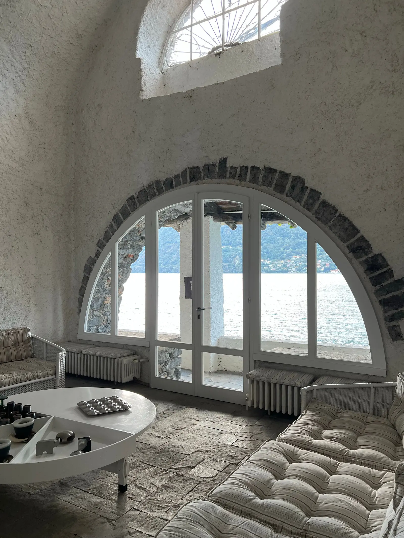 Sala con la finestra a mezzaluna aperta sul lago, divani bianchi e pavimento chiaro