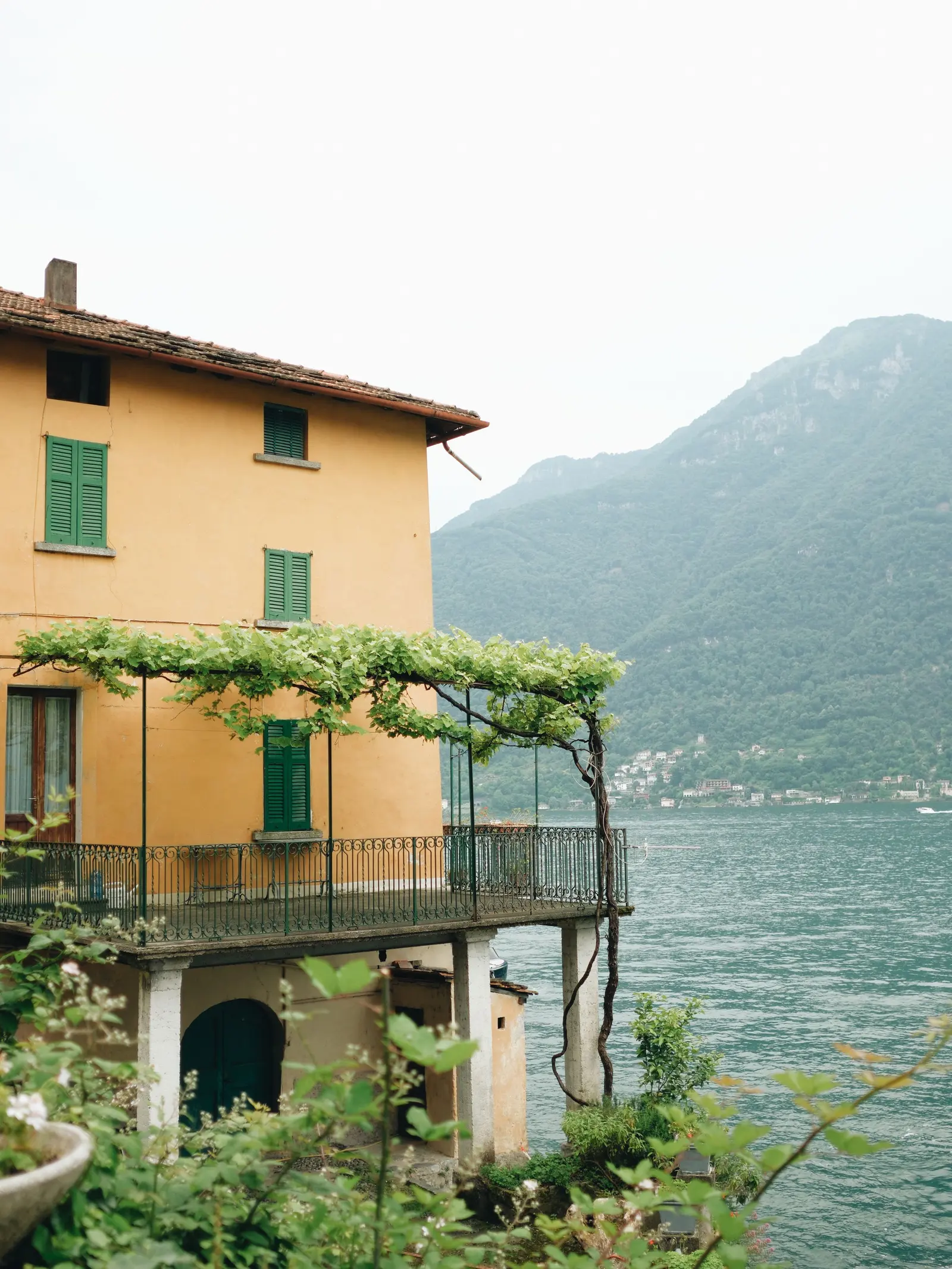 Casa color ocra con persiane verdi e un balcone coperto dalla vite, sopra l'acqua