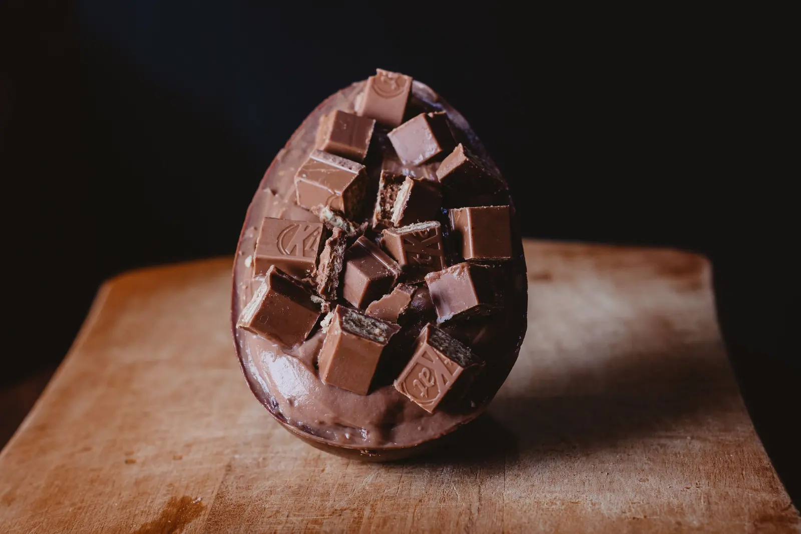 Un uovo di cioccolato con pezzi di cioccolato incastonati, su un tagliere di legno, fondo scuro