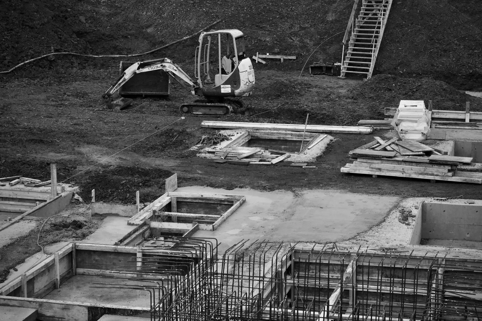 Fondazioni in costruzione: casseri, ferri d'armatura e un miniescavatore sullo sfondo, in bianco e nero
