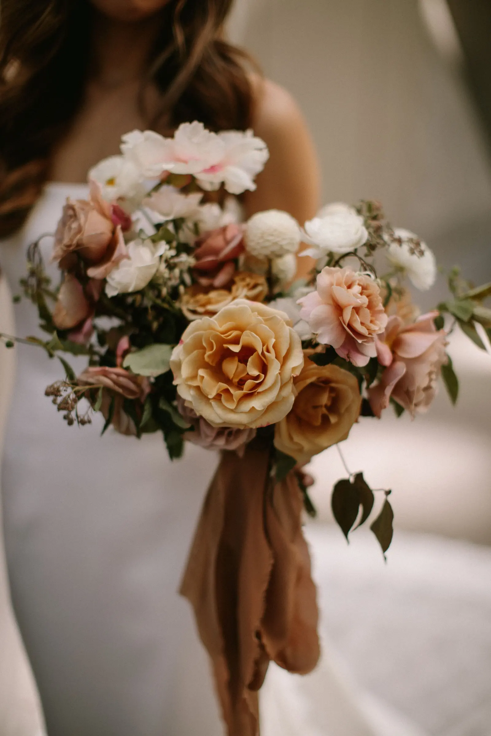 Una sposa tiene un bouquet di rose pesca e fiori misti legato con un nastro color ruggine