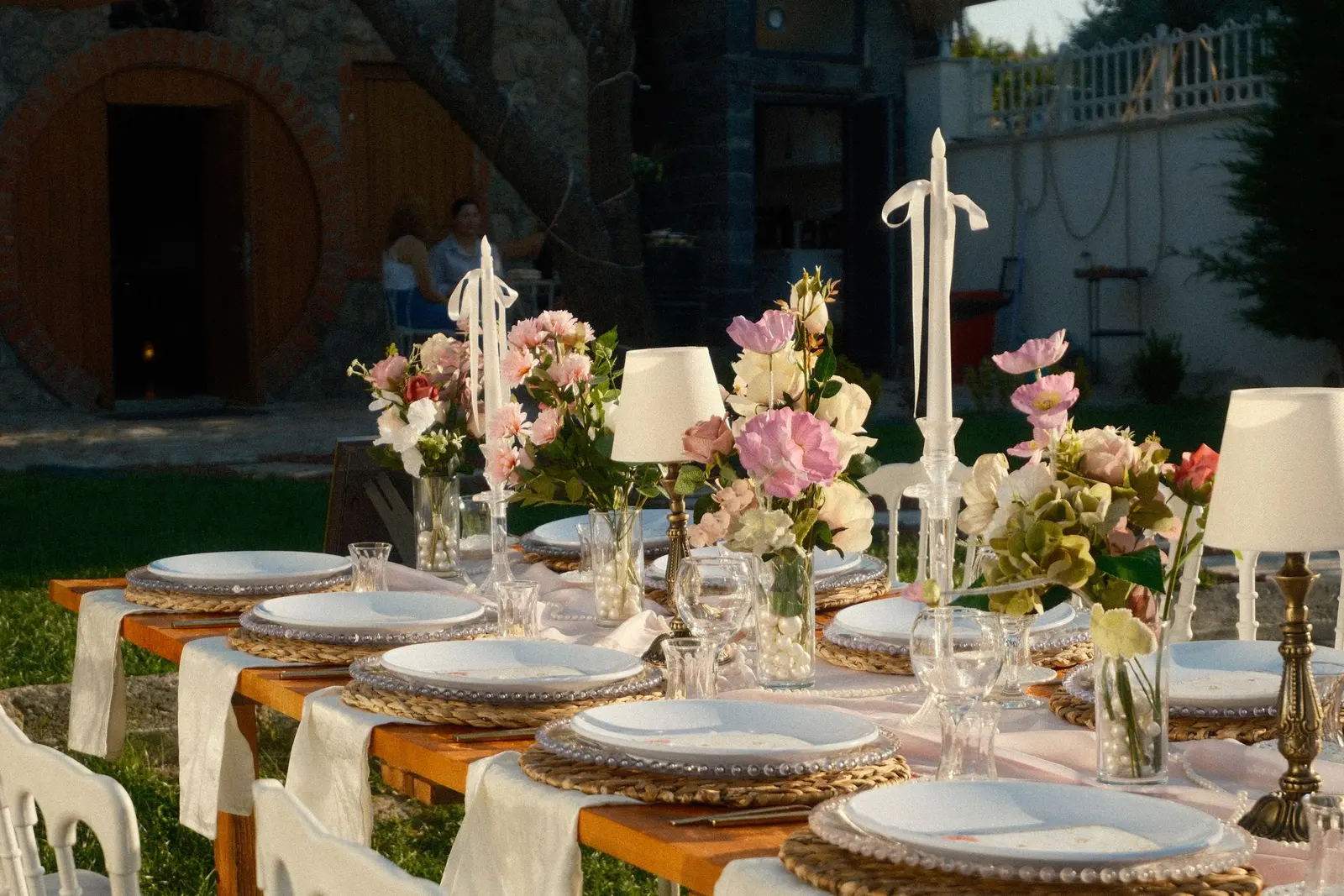 Tavola apparecchiata in giardino con composizioni basse di fiori rosa e pesca e candele