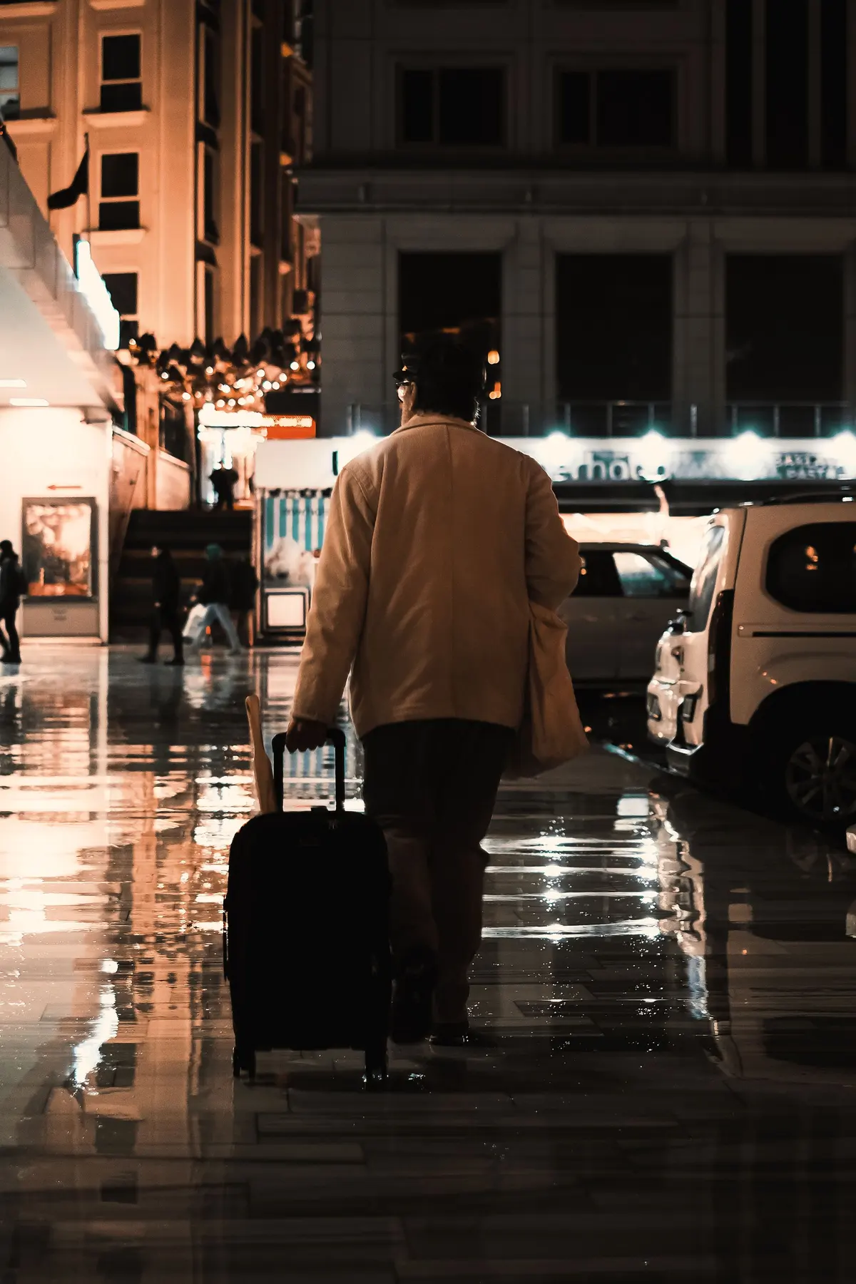 Una persona con il trolley su un marciapiede bagnato, la sera, tra le luci della città
