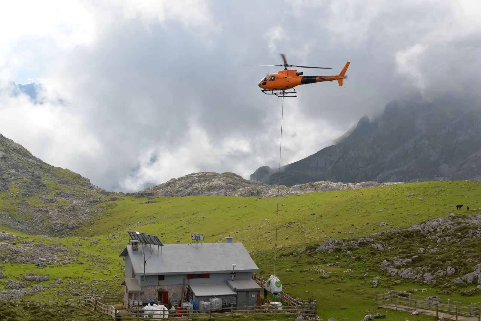 Un elicottero arancione porta un carico al gancio verso un rifugio con pannelli solari, tra le nuvole