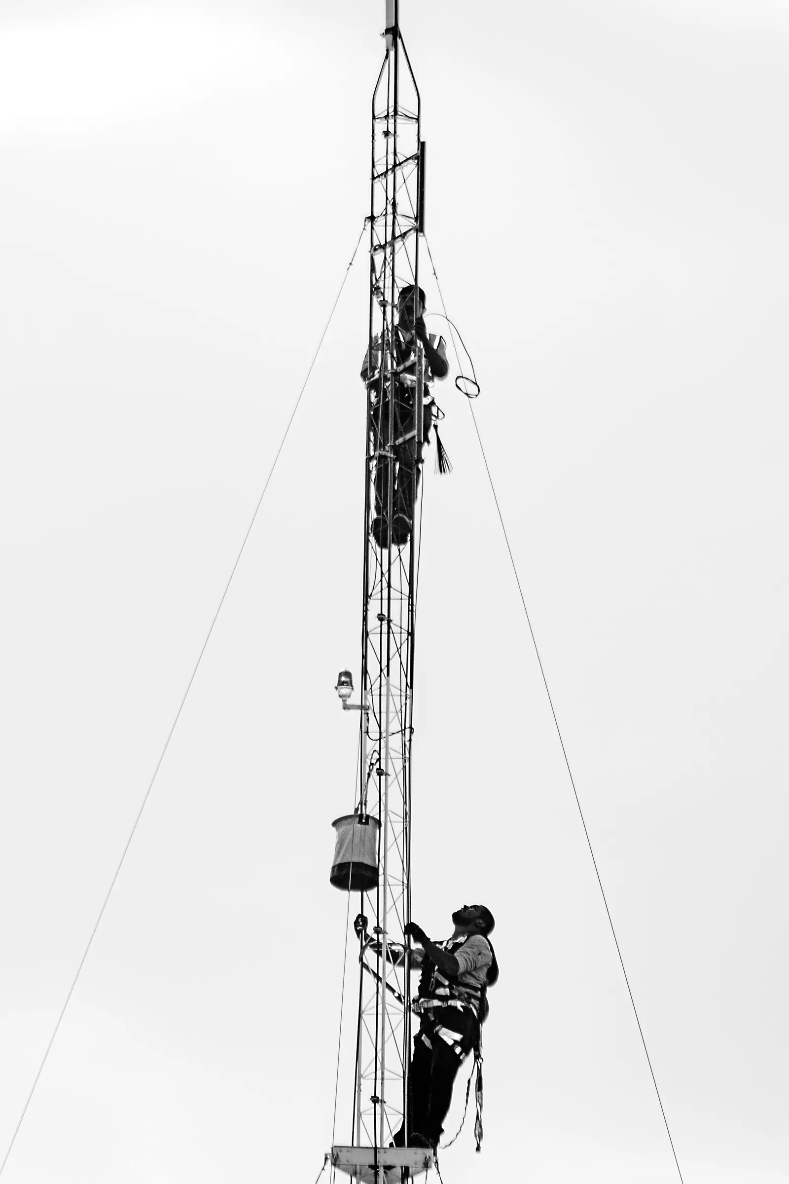 Due tecnici imbragati salgono un palo strallato, uno in cima e uno a metà, in bianco e nero