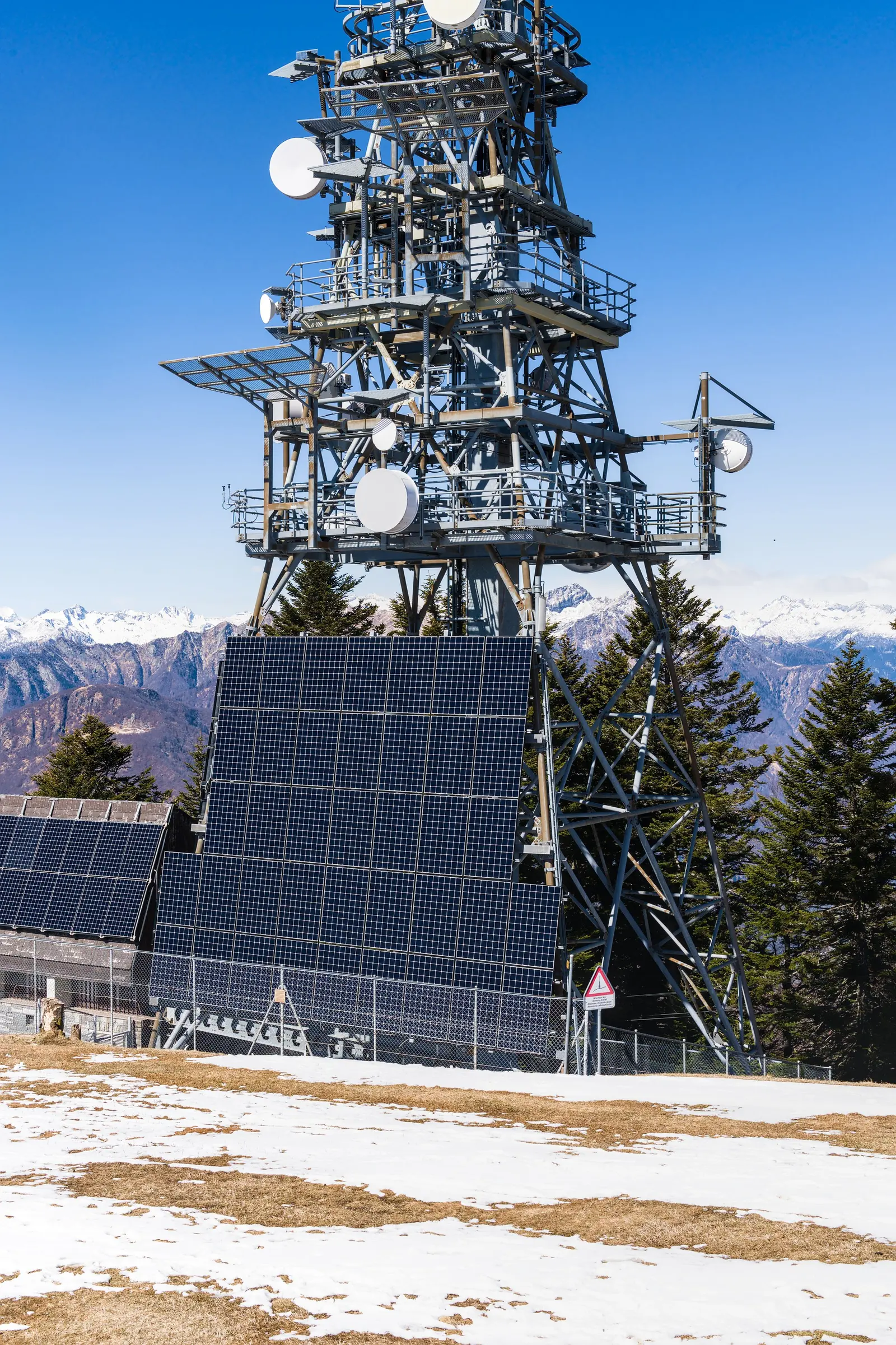 Un traliccio in quota con parabole e antenne, pannelli fotovoltaici alla base e neve sul prato