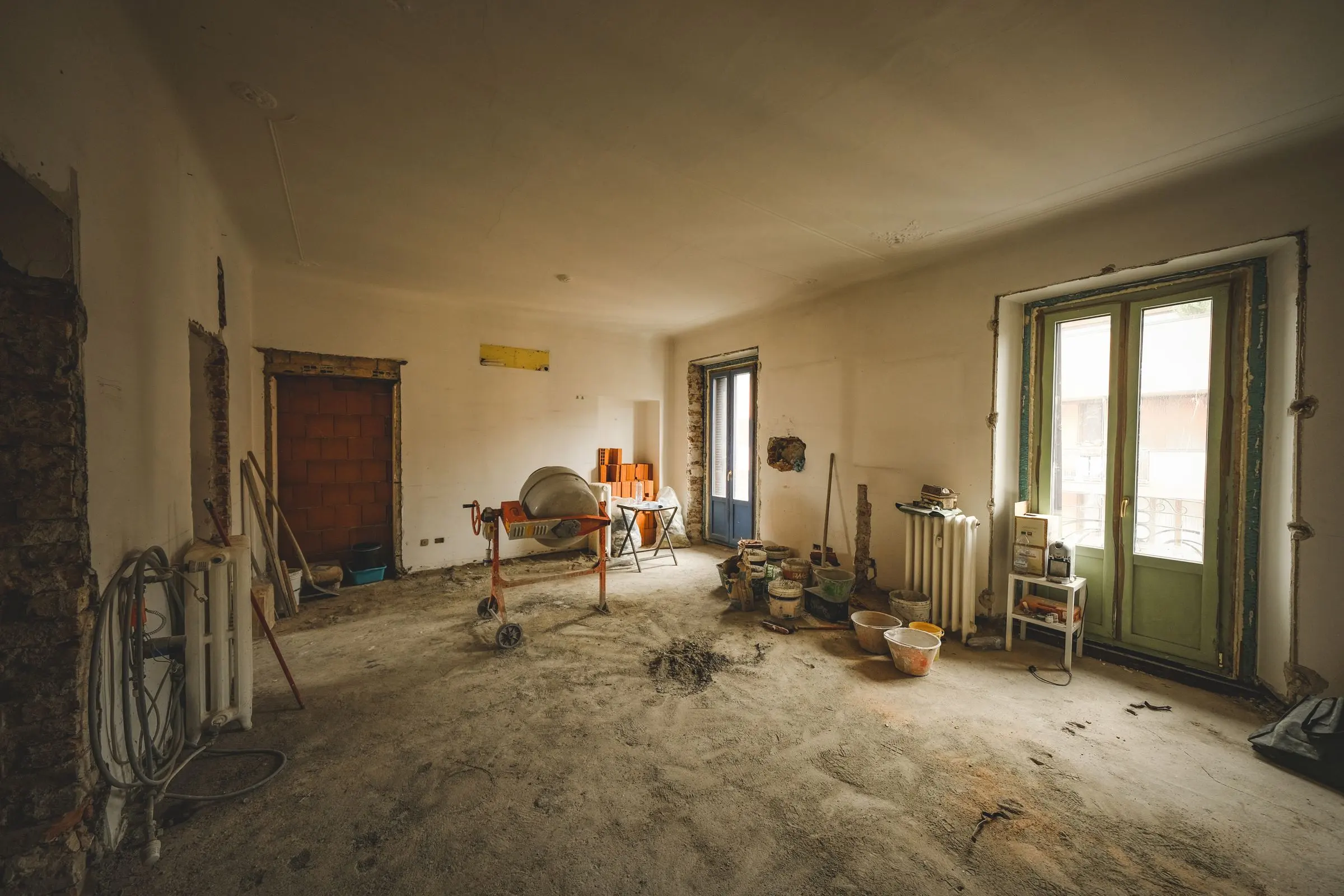 Prima: stanza grande in cantiere, radiatori staccati, attrezzi e sacchi sul massetto, portefinestre in fondo