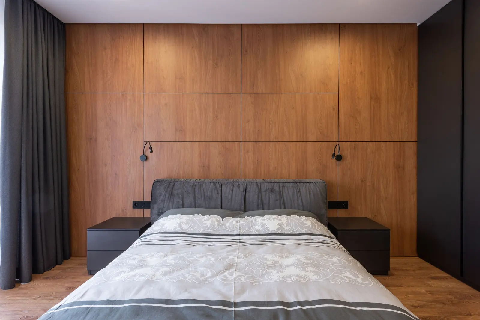 Camera con letto imbottito grigio davanti a una parete di pannelli in legno con due lampade da lettura