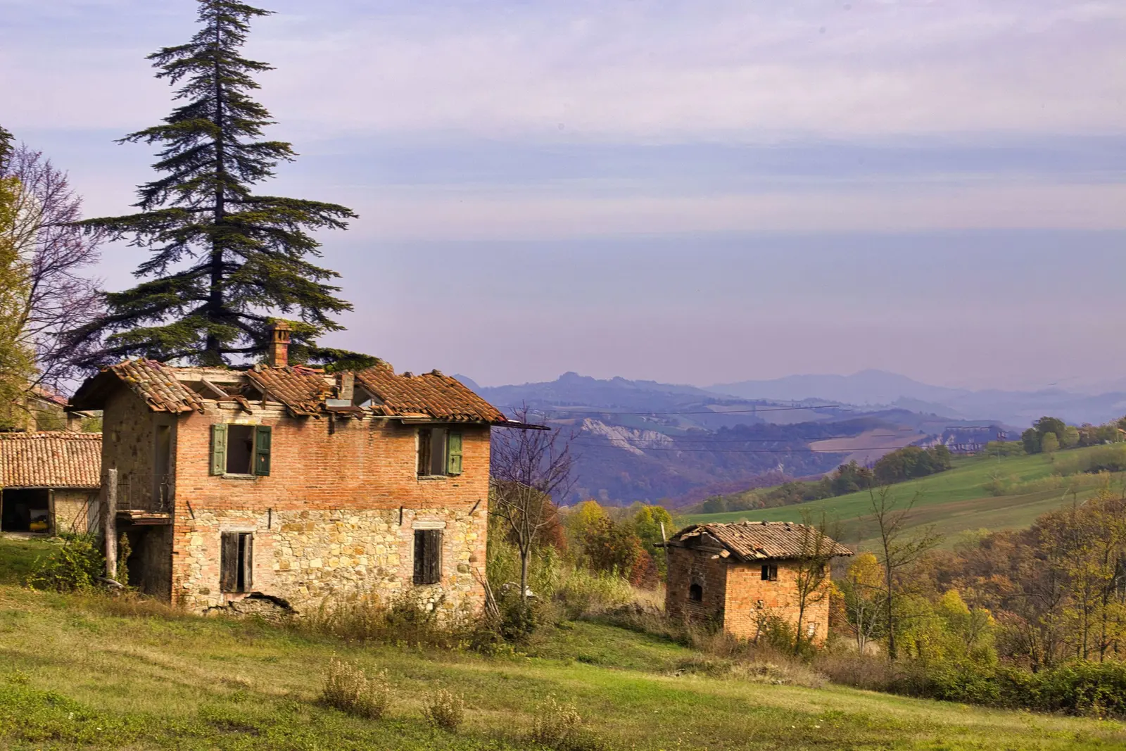 Un casolare abbandonato in mattoni tra le colline, con un grande cedro accanto e il cielo del tramonto