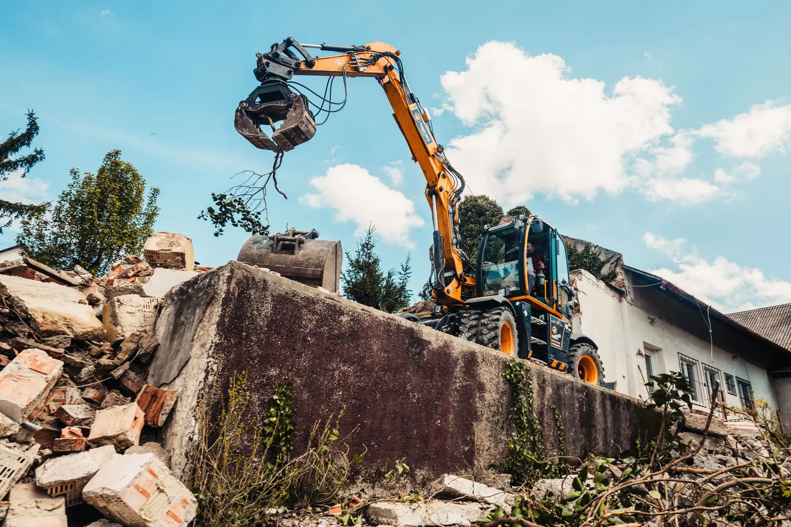 Un escavatore con la pinza da demolizione sopra un muro di cemento, con le macerie davanti e una casa bianca dietro