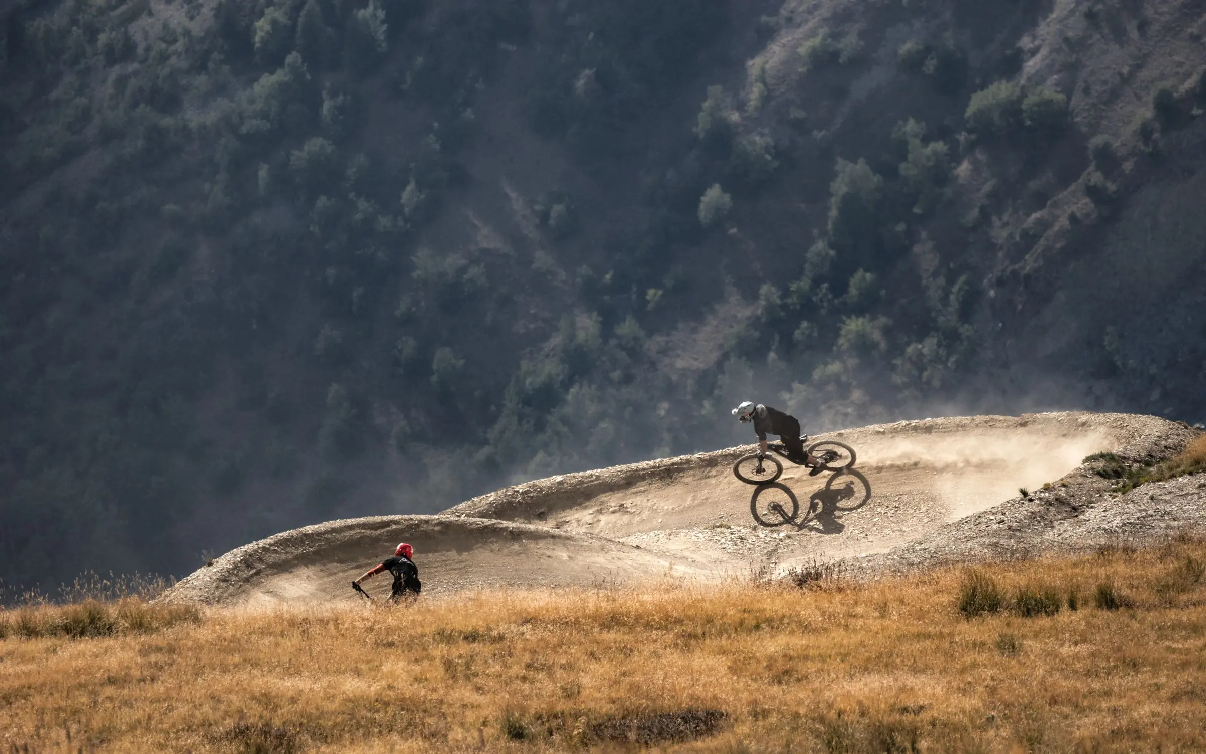 Due rider in discesa su curve paraboliche polverose in un pascolo d'alta quota, dietro un versante scuro