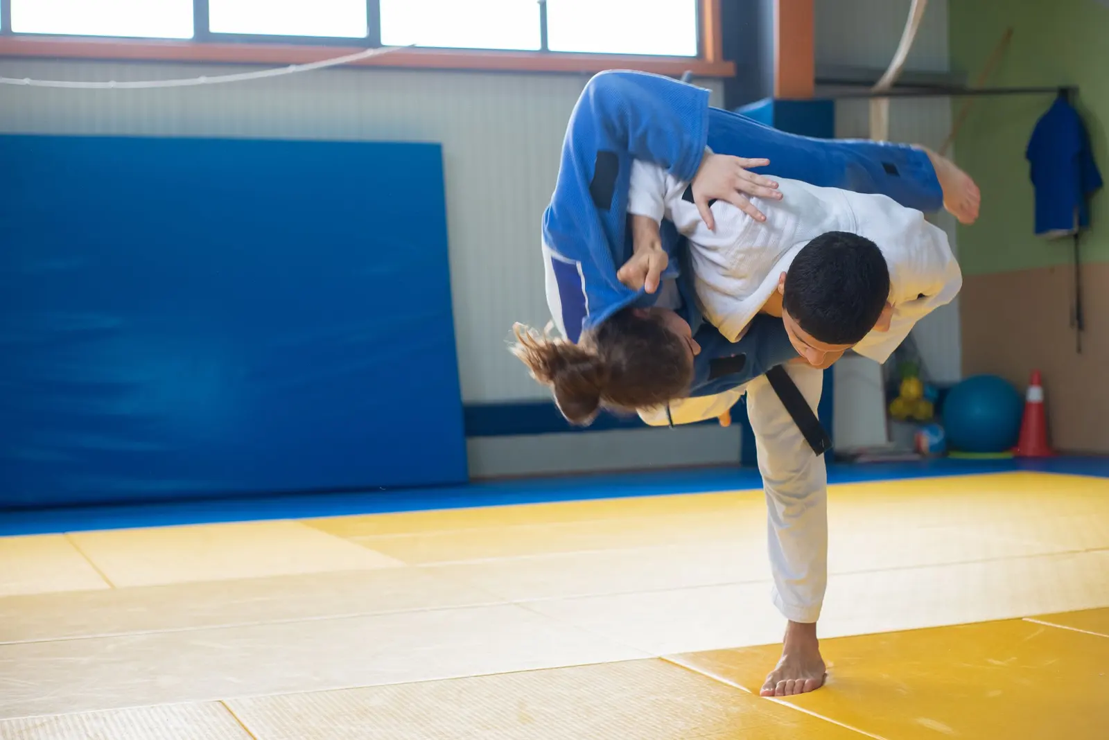 Proiezione di judo sul tatami giallo: chi esegue in judogi bianco, chi cade in judogi blu