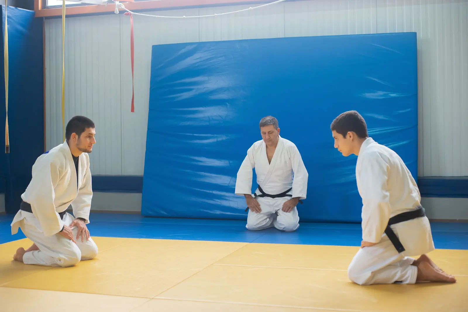 Tre judoka in ginocchio sul tatami, l'insegnante in fondo davanti al muro blu