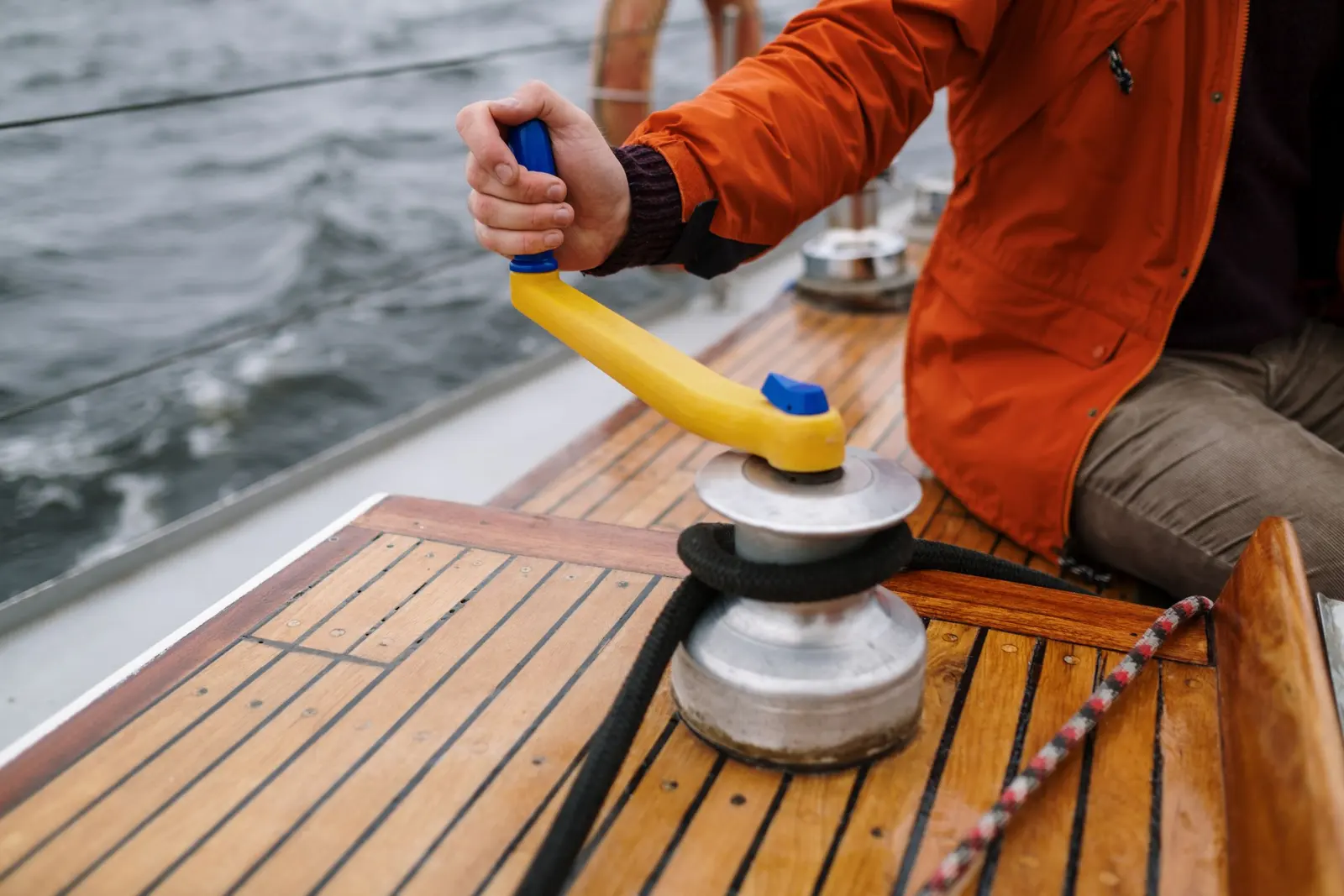 Mano sulla manovella gialla di un winch, con la coperta in teak e il mare dietro