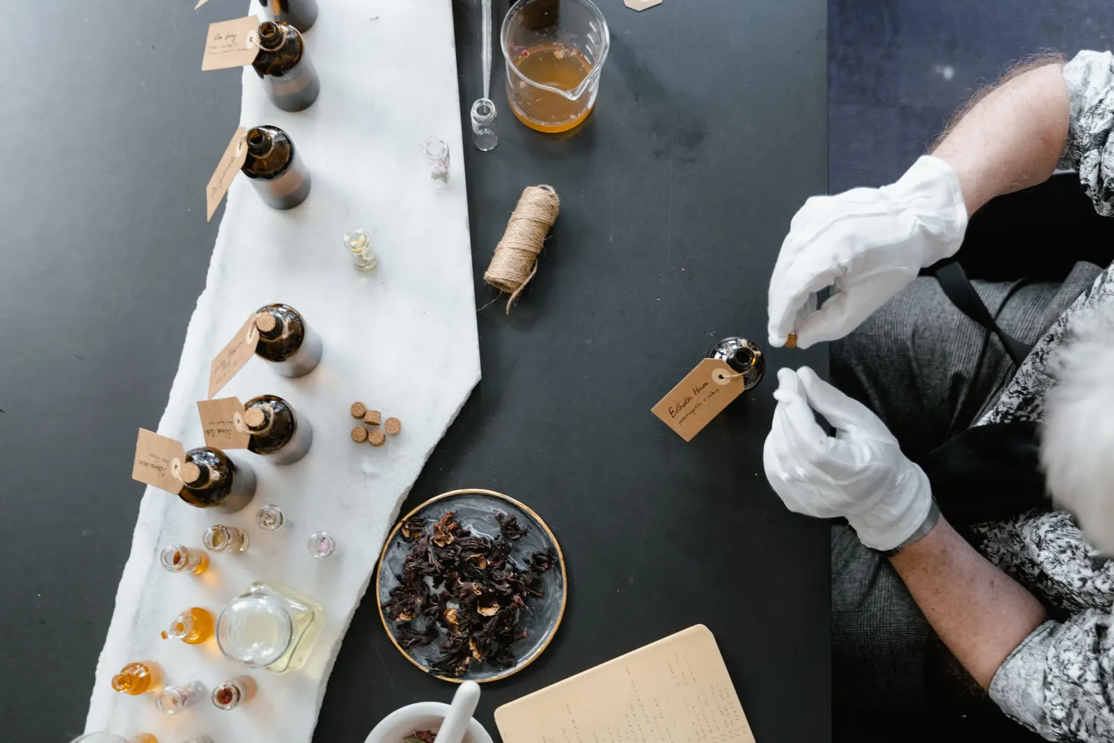 Il banco nero del laboratorio visto dall'alto: guanti, flaconi, fiori secchi e cartellini scritti a mano