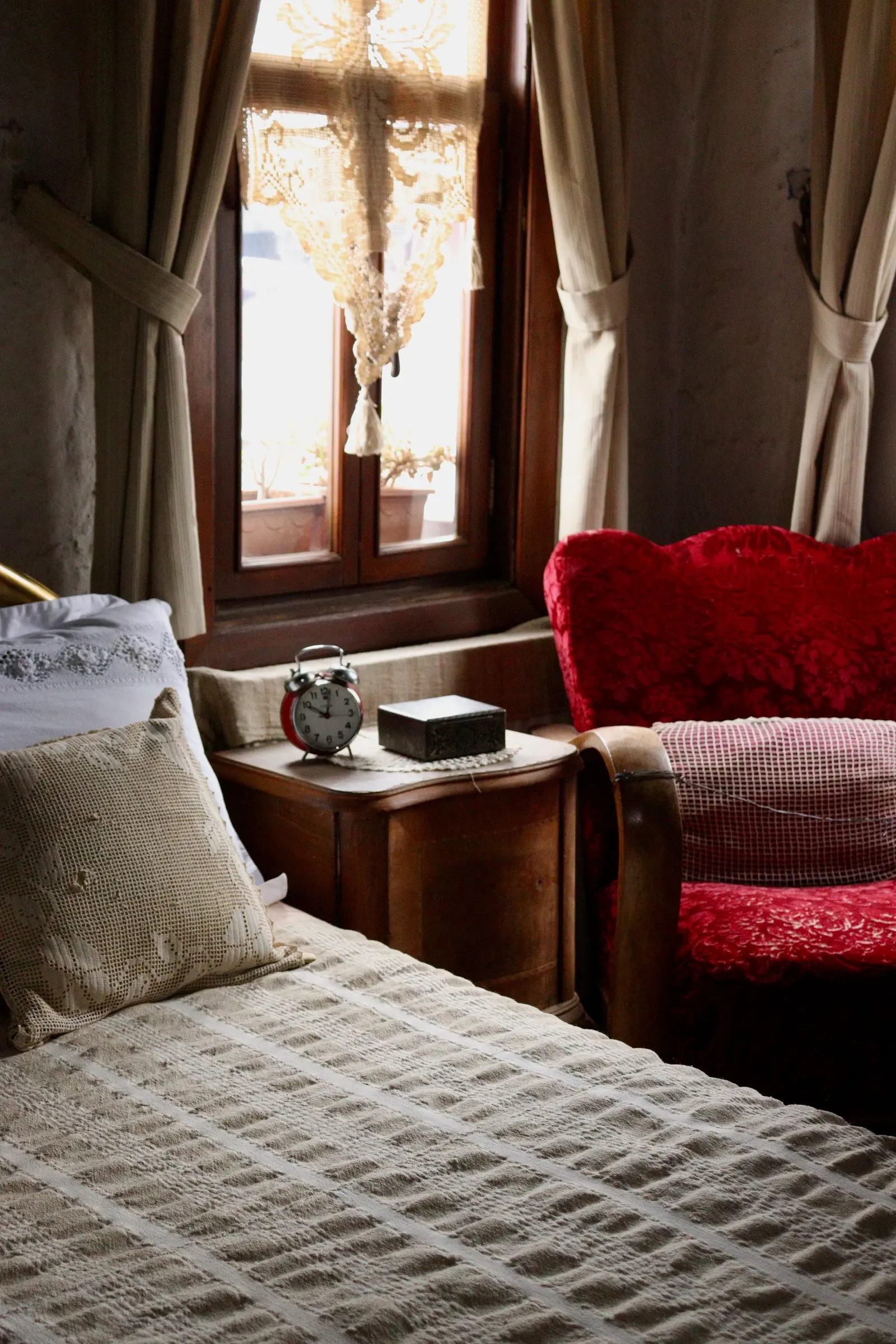Angolo di camera con poltrona di velluto rosso, comò antico e letto con copriletto chiaro