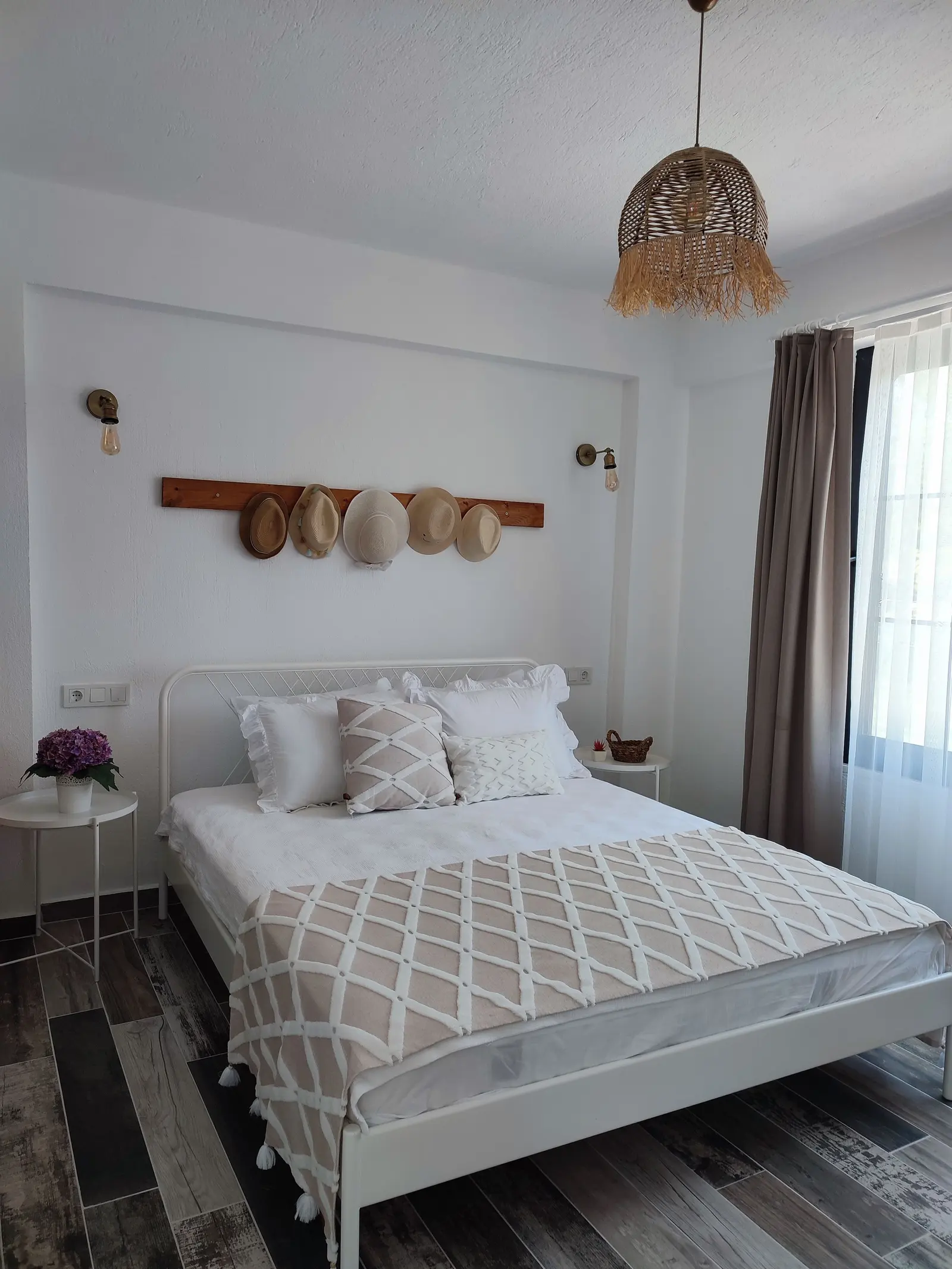 Camera bianca con letto matrimoniale, coperta a rombi, lampada di paglia e cappelli appesi al muro