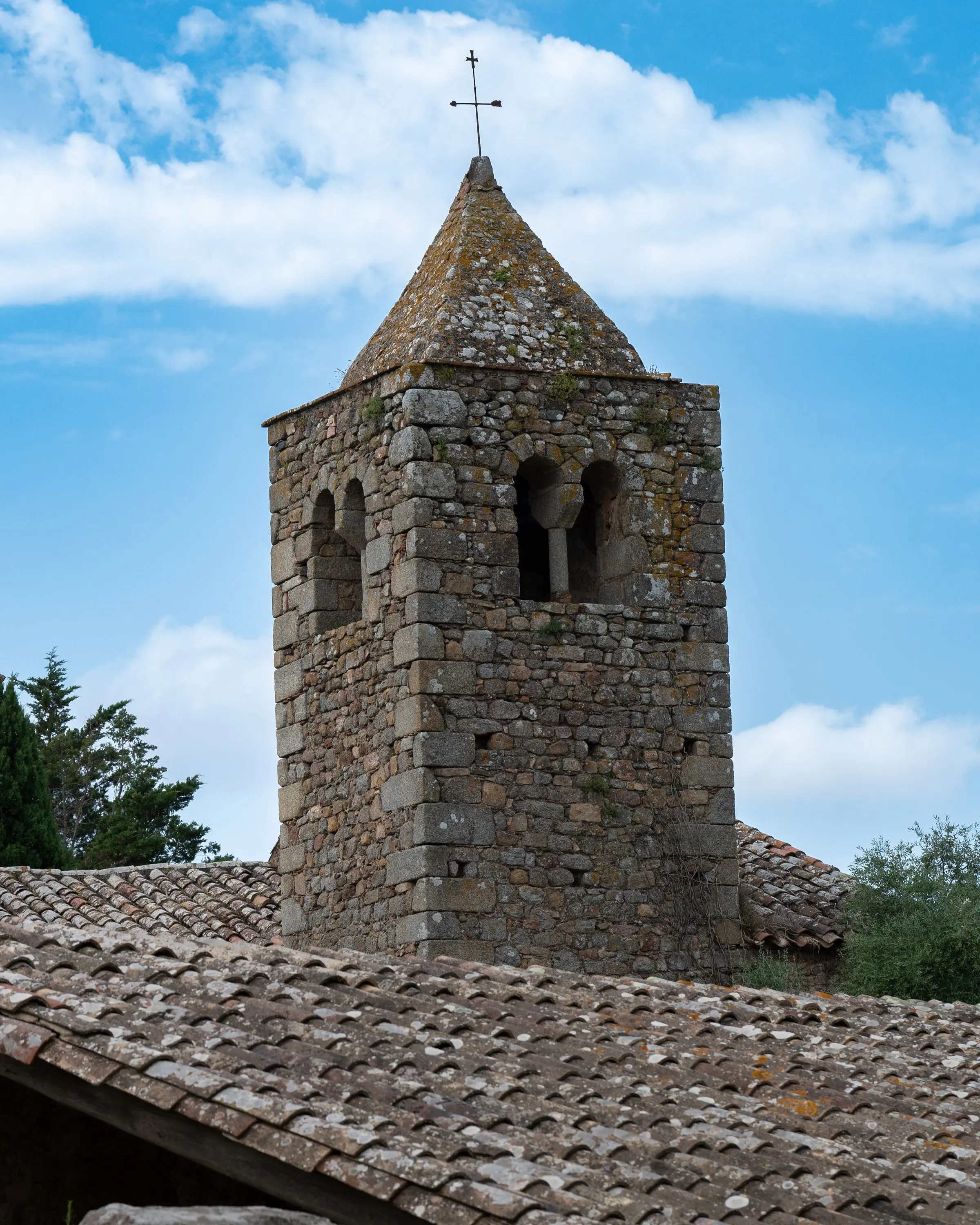 Campanile in pietra con bifore e cuspide, sopra un tetto in coppi, contro un cielo azzurro