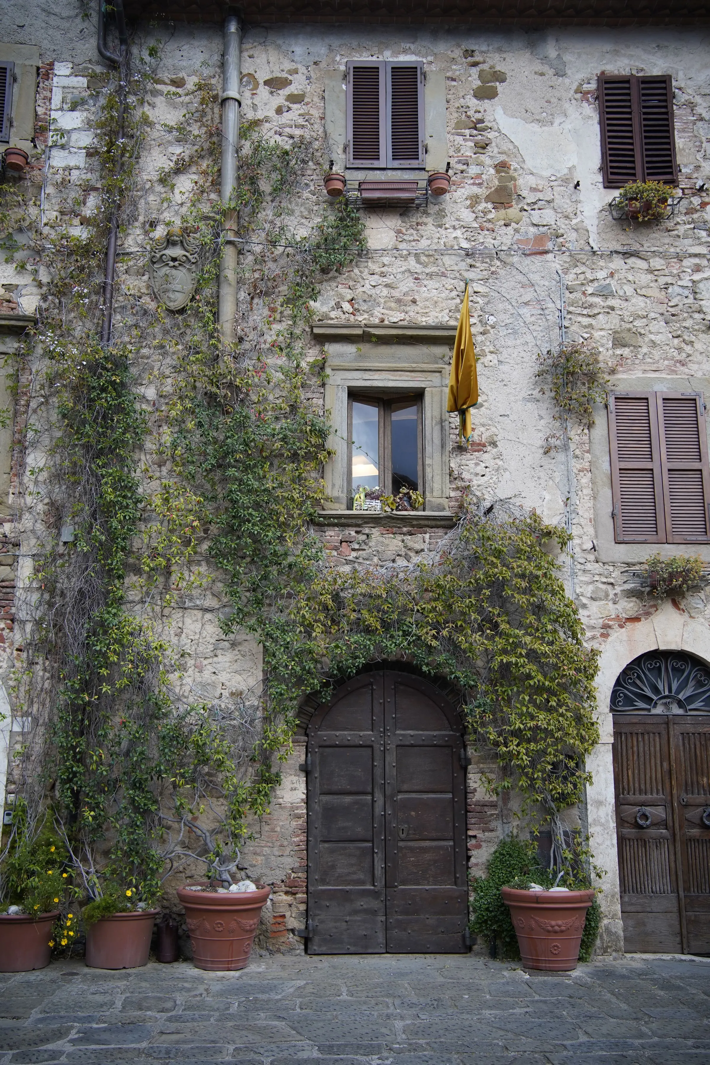 Facciata in pietra di una casa di borgo con edera, portone ad arco in legno e vasi di piante