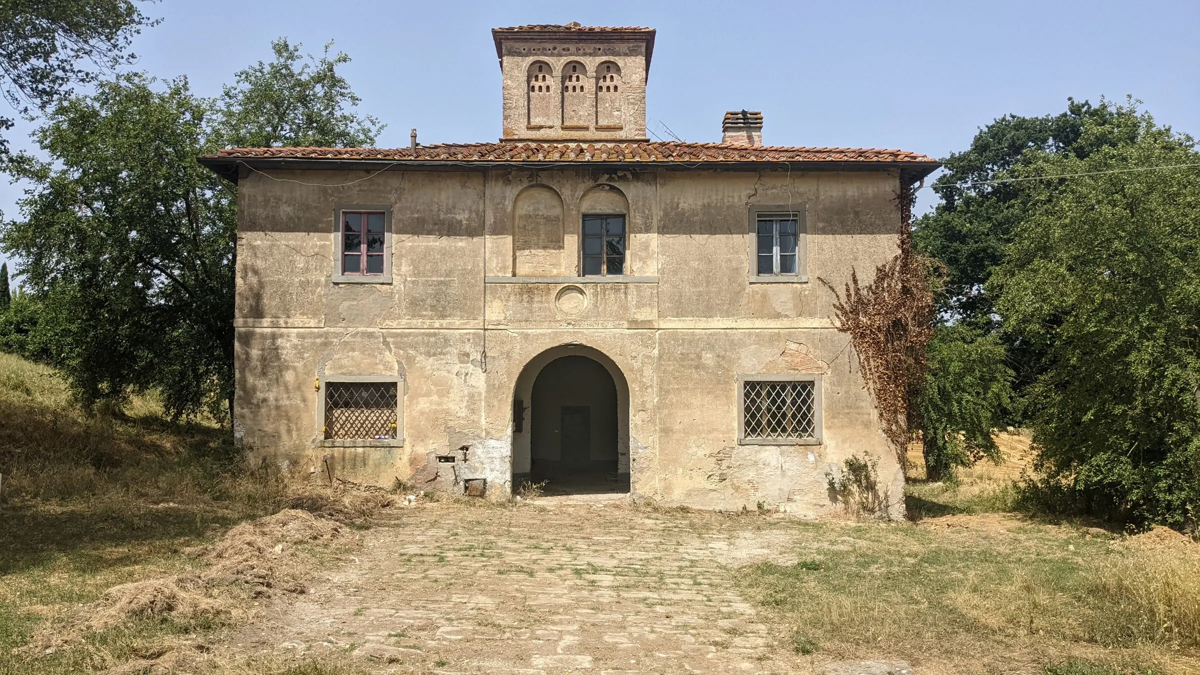 Villa di campagna su due piani con altana a tre archi, intonaco consumato e portale ad arco, tra alberi e prato secco