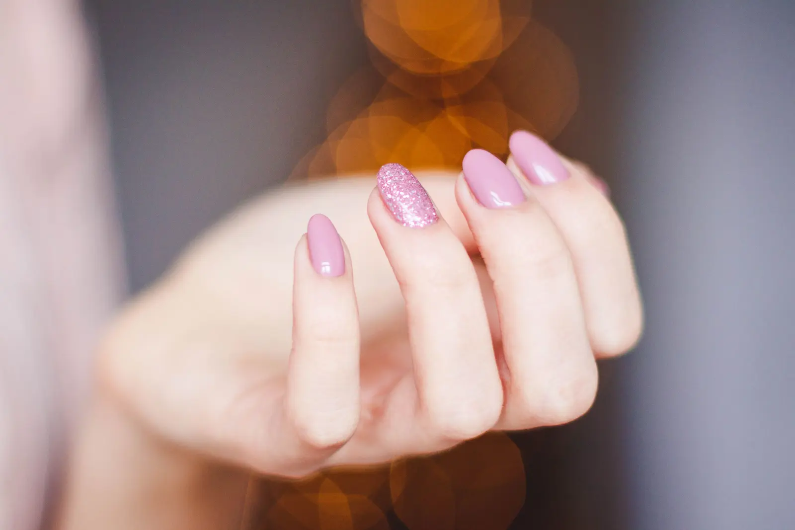 Unghie rosa a mandorla, una con glitter, con luci calde sfocate