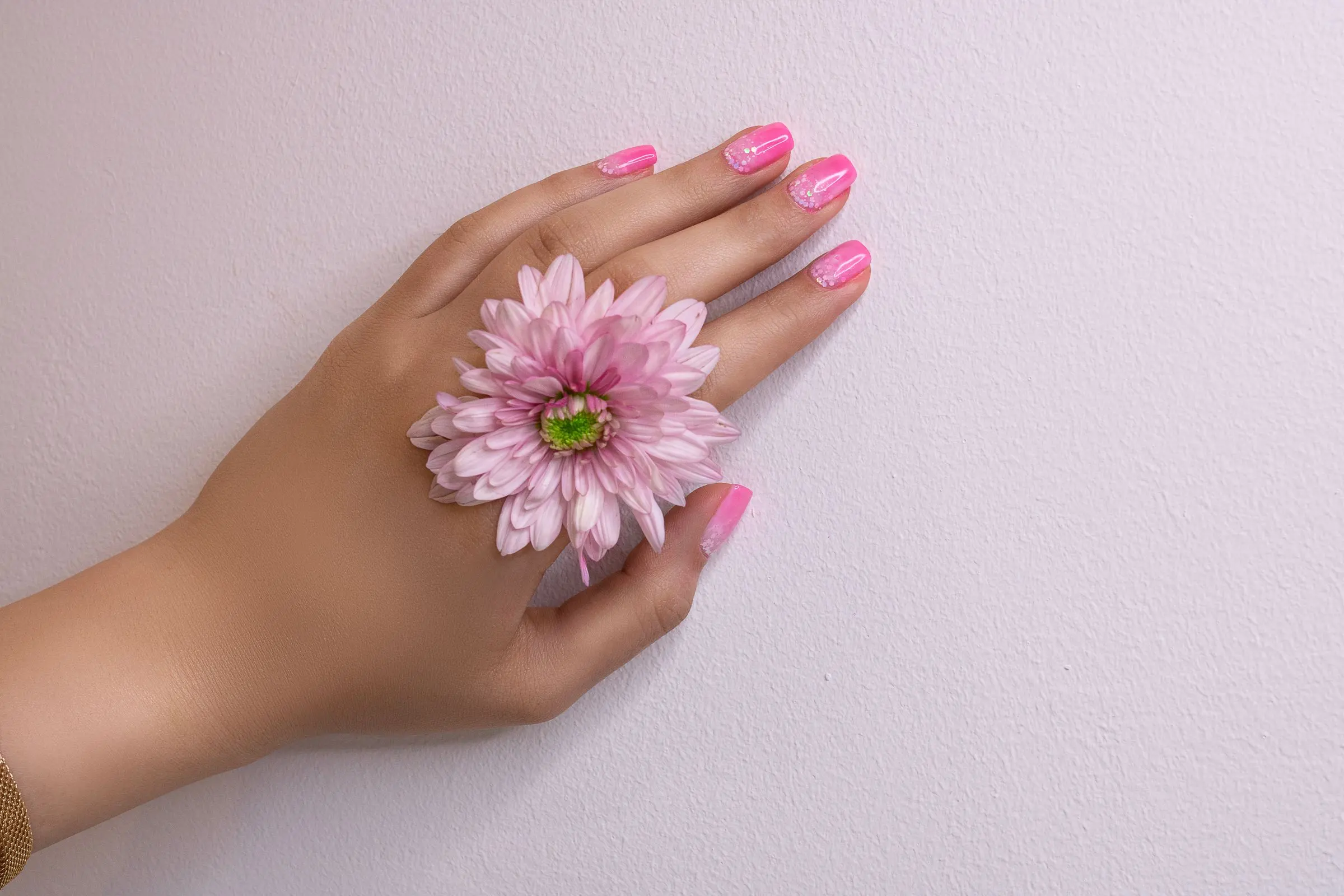 Mano con smalto rosa e un fiore rosa appoggiato tra le dita, su fondo lilla