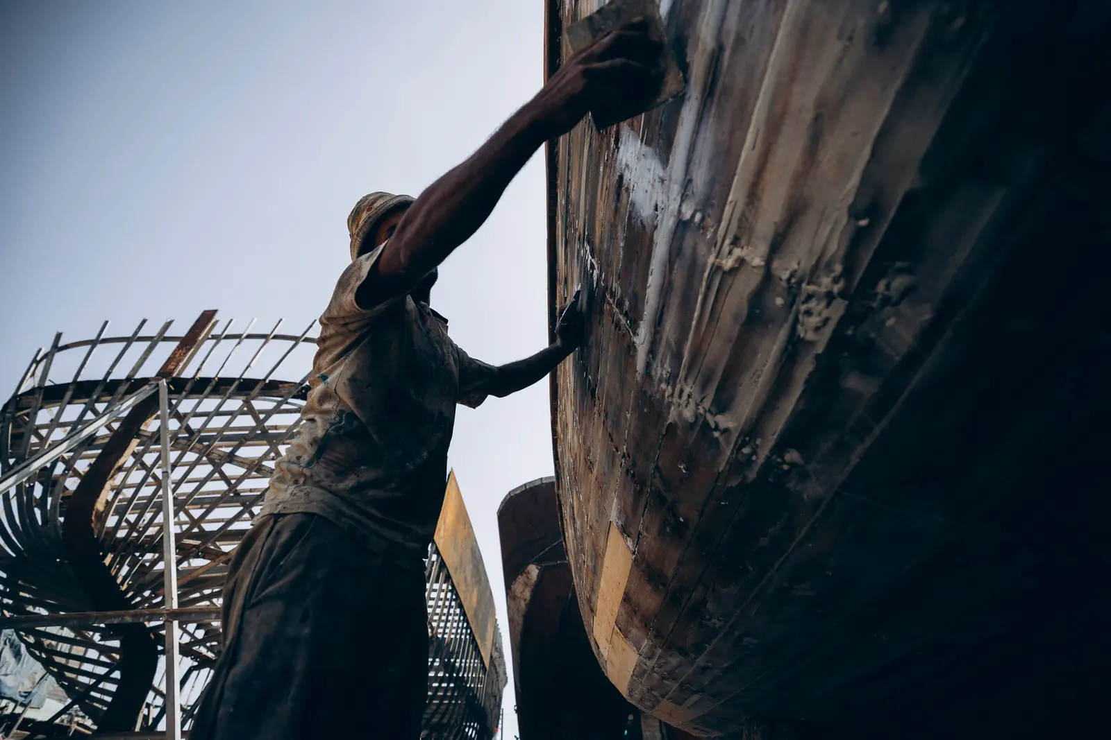 Un uomo lavora con le mani sul fianco di uno scafo in legno, in un cantiere all'aperto