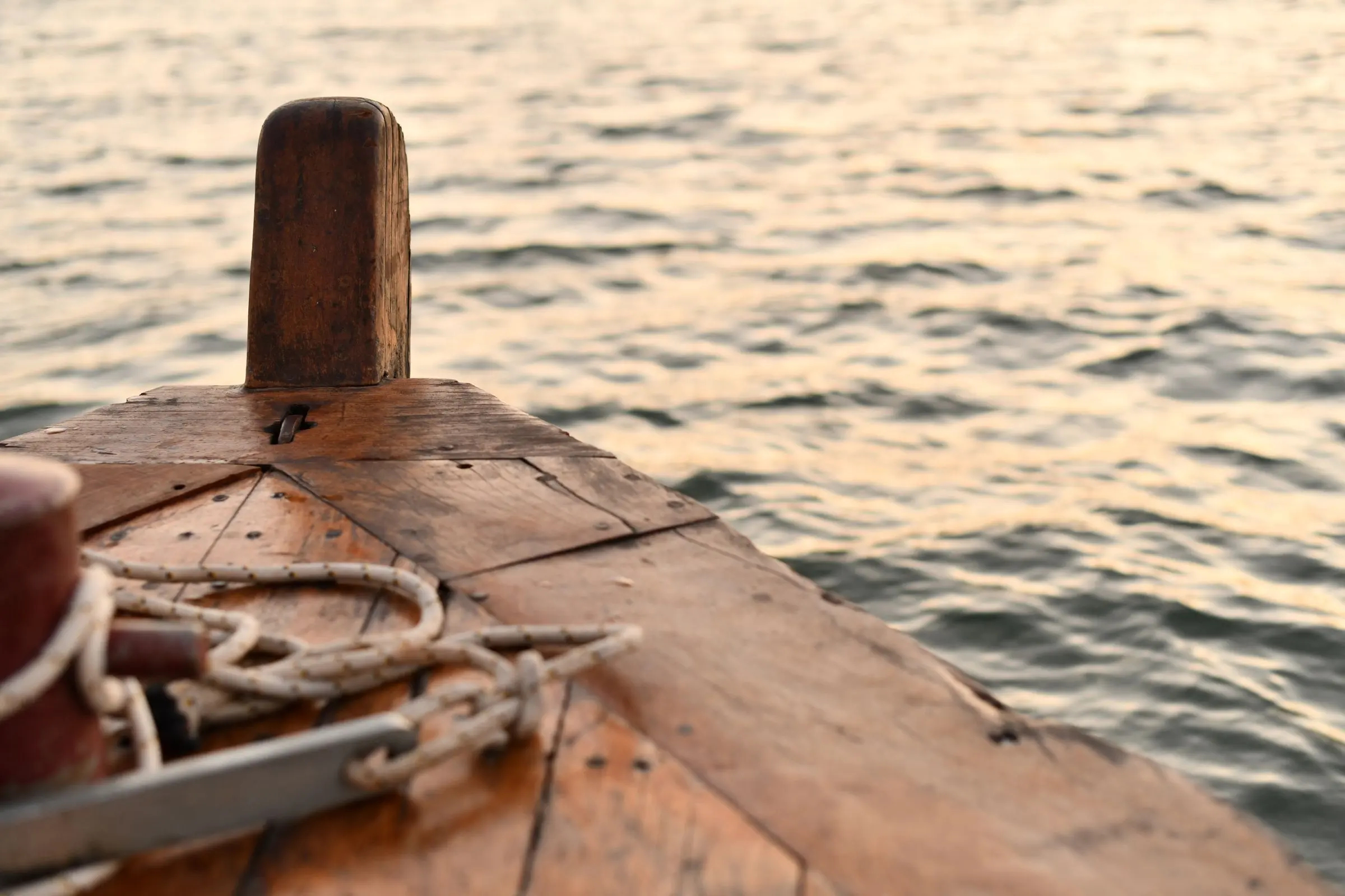 Prua di una barca in legno con la bitta e una cima avvolta, davanti al mare dorato del tramonto