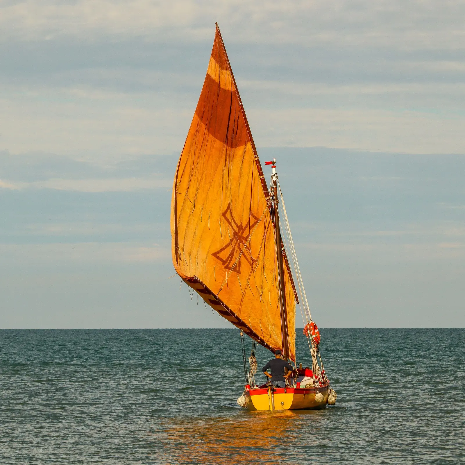 Una piccola barca tradizionale naviga con la vela al terzo color ocra su un mare calmo