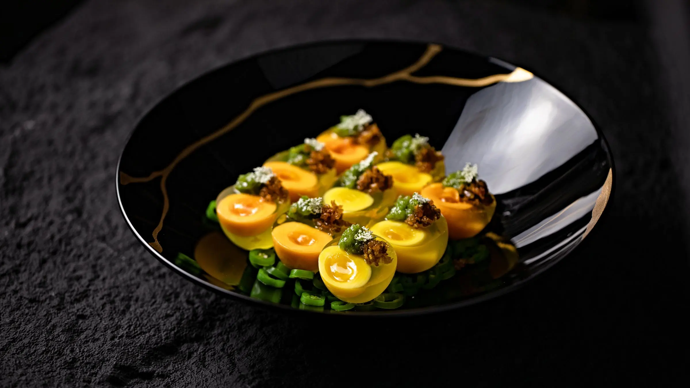 Mezze uova marinate con salsa verde e pane sbriciolato, su fagiolini, in un piatto nero con venature dorate