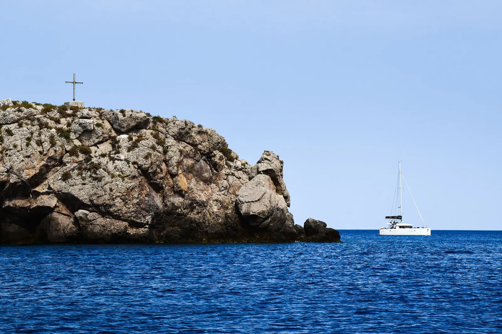 Catamarano alla fonda sul mare blu, accanto a uno scoglio con una croce in cima
