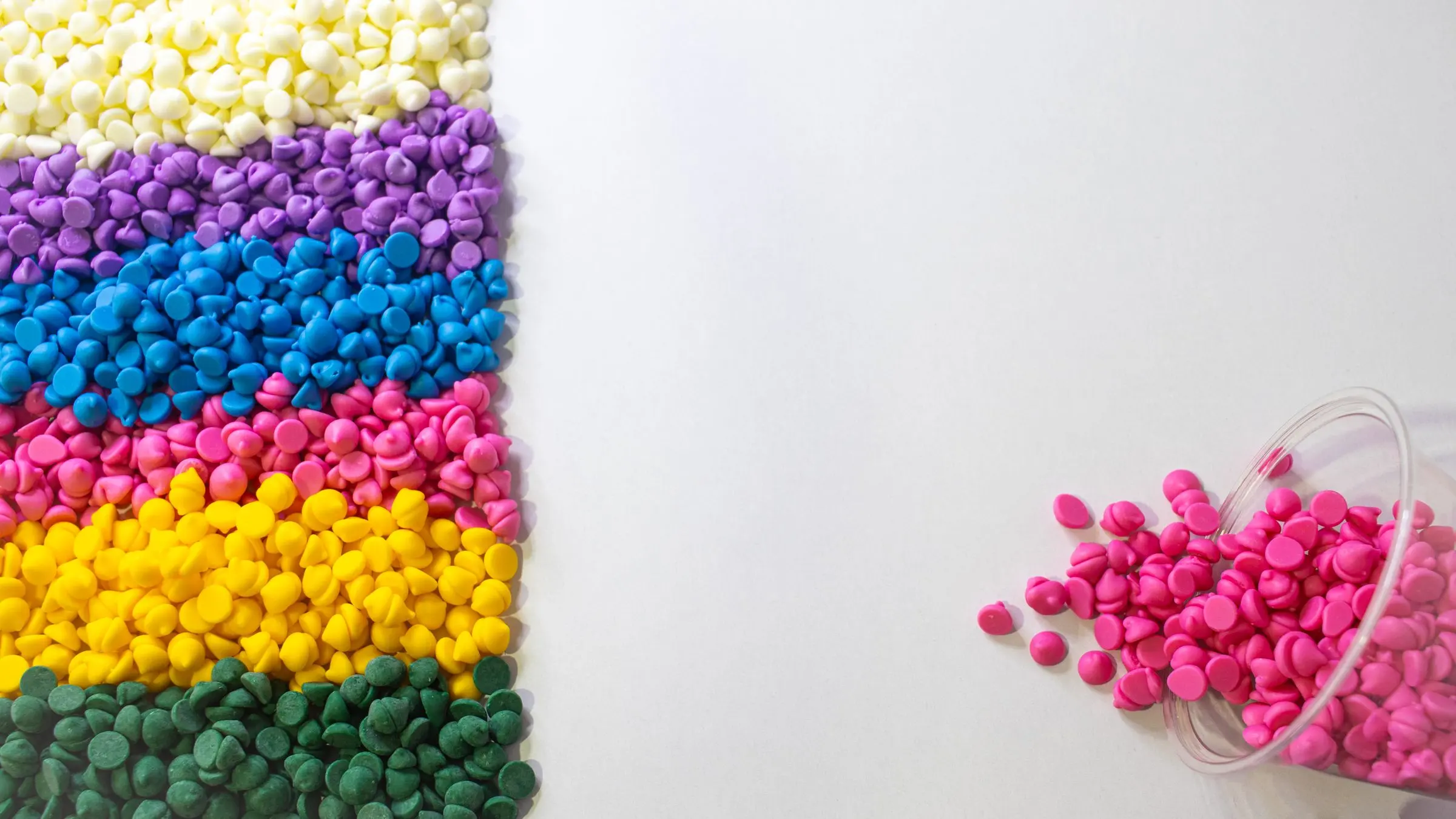 Granuli di plastica colorati disposti a strisce: bianco, viola, blu, magenta, giallo e verde; a destra un bicchiere rovesciato di granuli magenta