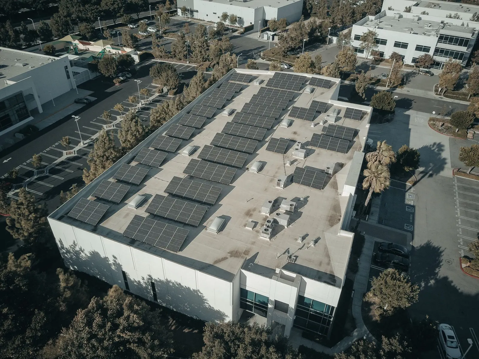 Edificio con tetto piano visto dal drone: file di moduli inclinati tra lucernari e macchine, palme intorno