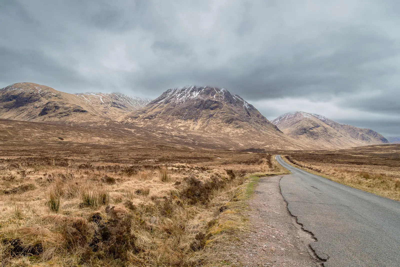 Una strada stretta che attraversa la brughiera delle Highlands verso montagne con la neve in cima
