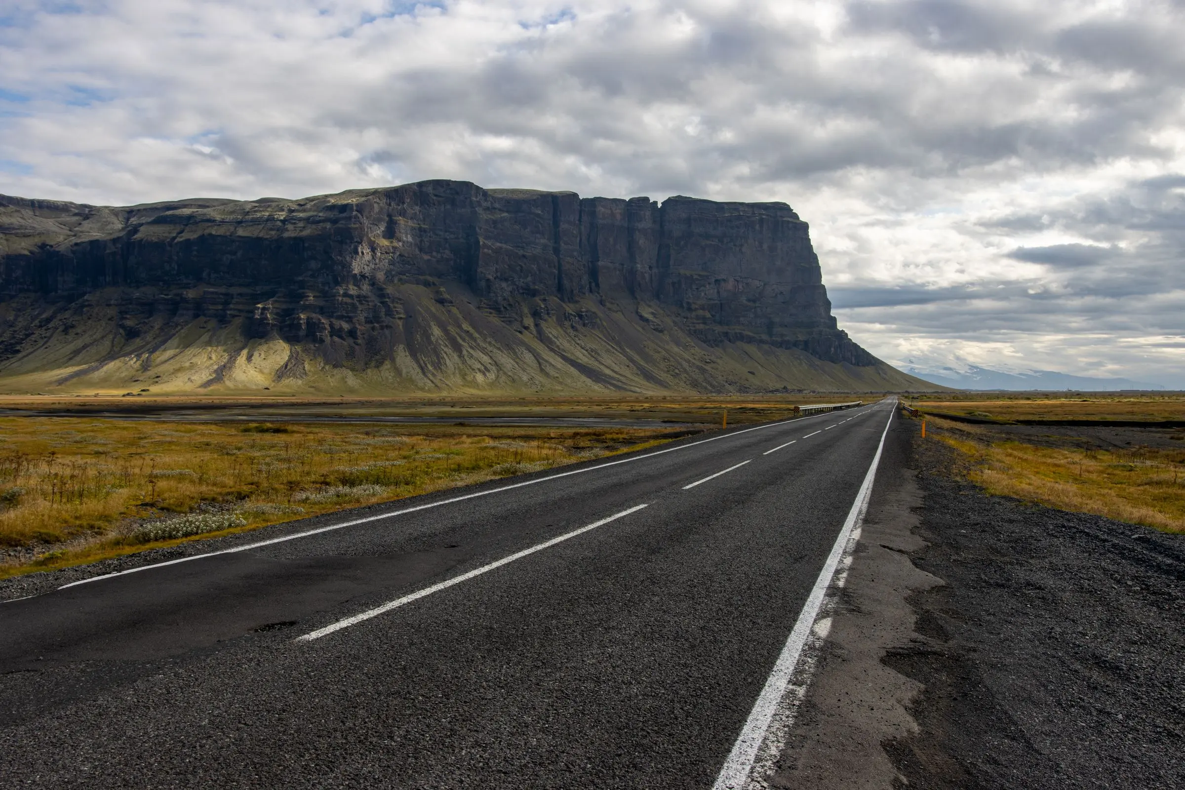 Una strada islandese dritta e vuota che corre verso una falesia a tavolato, sotto un cielo grigio
