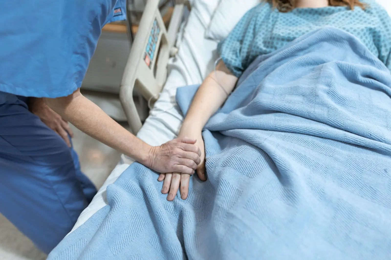 Un'infermiera tiene la mano di una paziente a letto, sotto una coperta azzurra