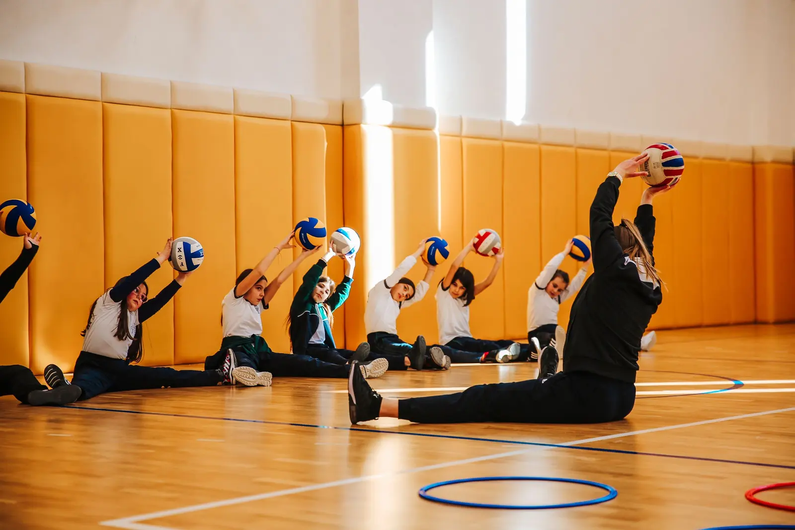 Bambine e bambini seduti in palestra fanno stretching con il pallone sopra la testa, con l'istruttrice