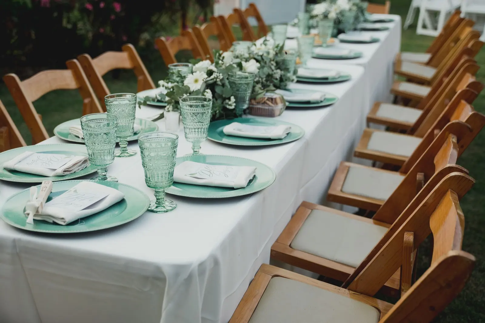 Tavolo di nozze apparecchiato con tovaglia bianca, piatti verde acqua, calici lavorati e fiori bianchi, sedie in legno